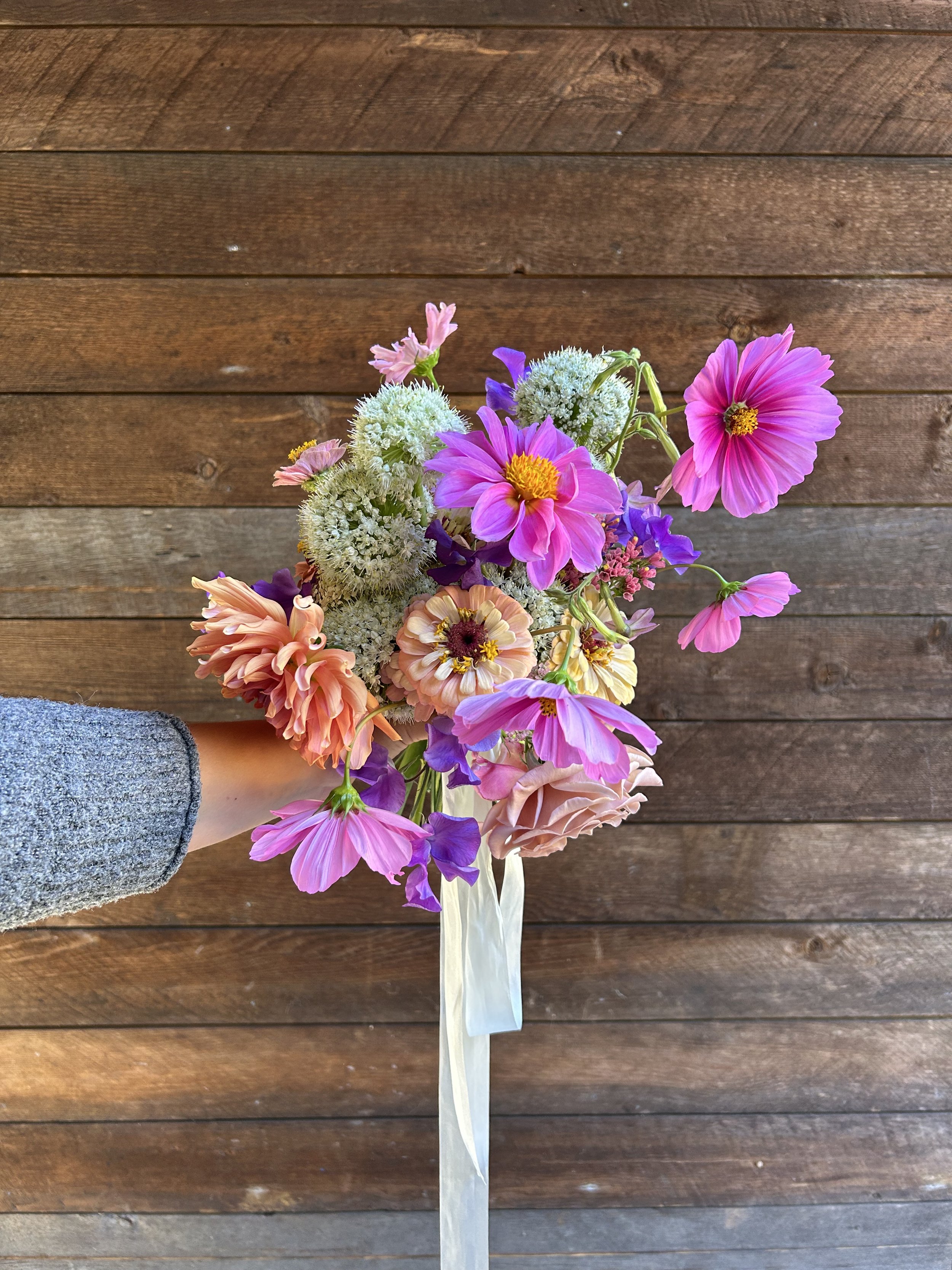 A hand holding a bridal bouquet of colorful flowers in front of a wooden background.