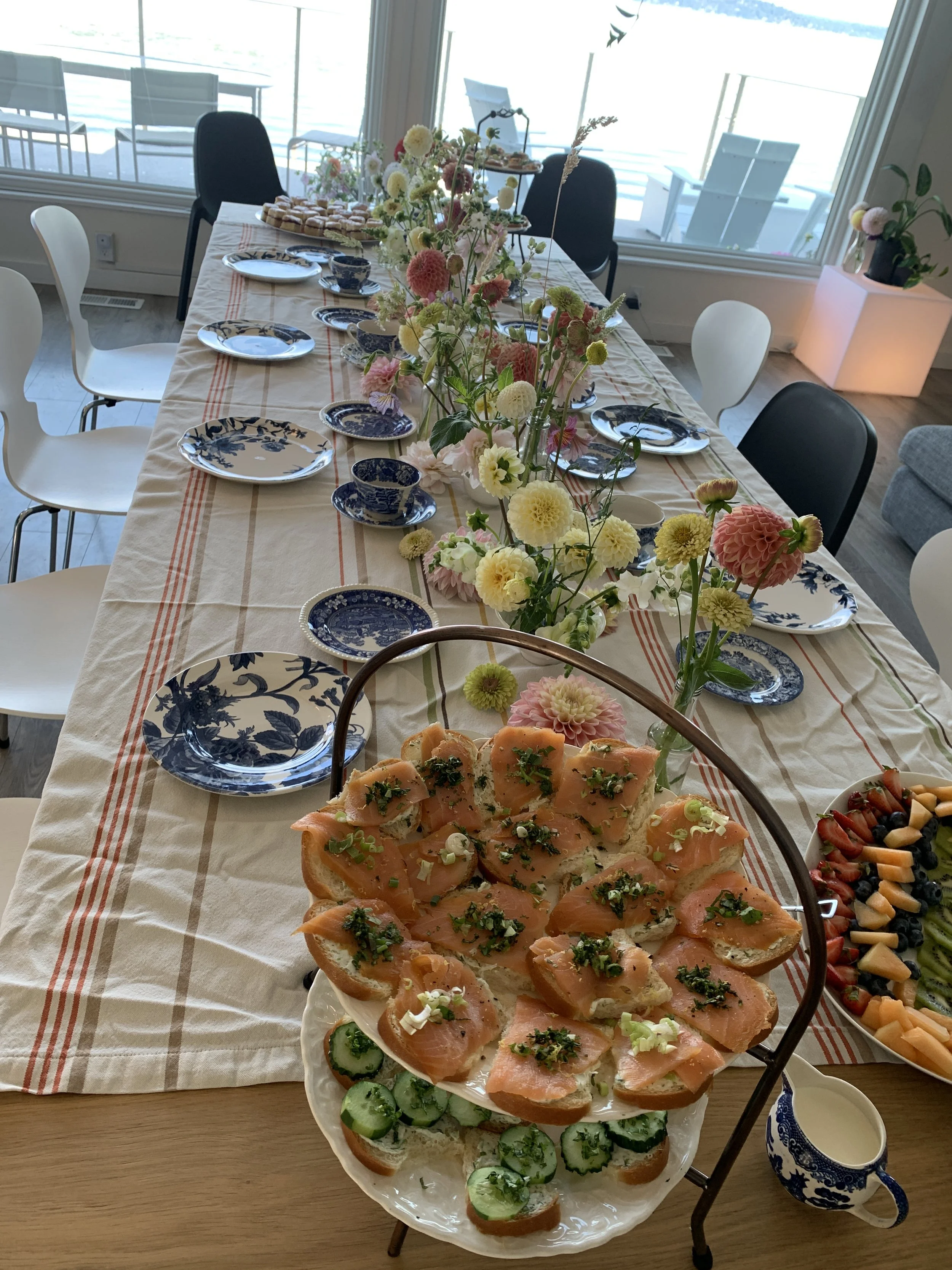 A long dining table set for a gathering with a beige tablecloth with red and beige stripes, decorated with a central arrangement of pink, white, and cream flowers. The table has blue and white plates, teacups, and a variety of appetizers including sm