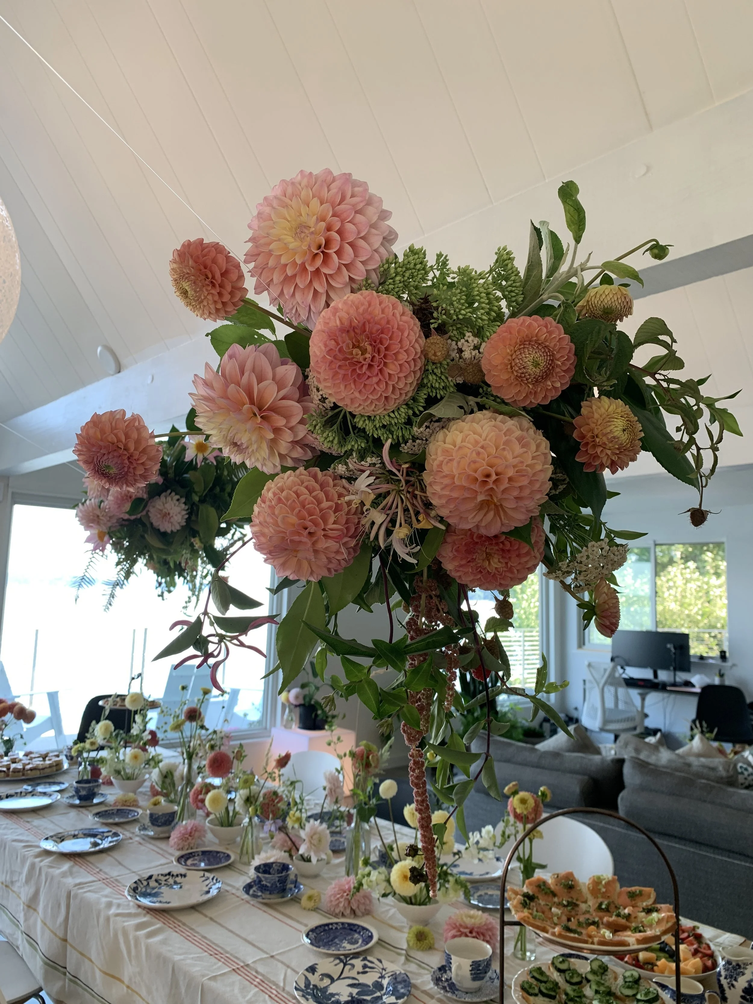 A large floral centerpiece with pink dahlias and green foliage displayed above a dining table set with blue and white china, surrounded by smaller flower arrangements and food.