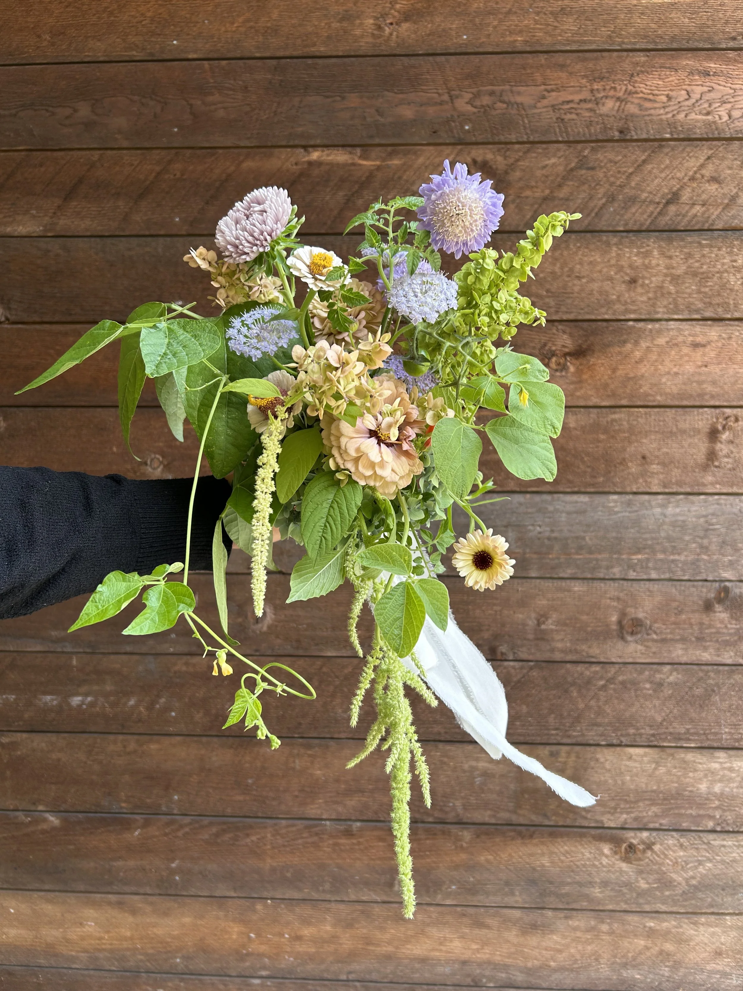 Hand holding a bridal bouquet of mixed flowers with purple, white, and yellow blooms against a wooden background.