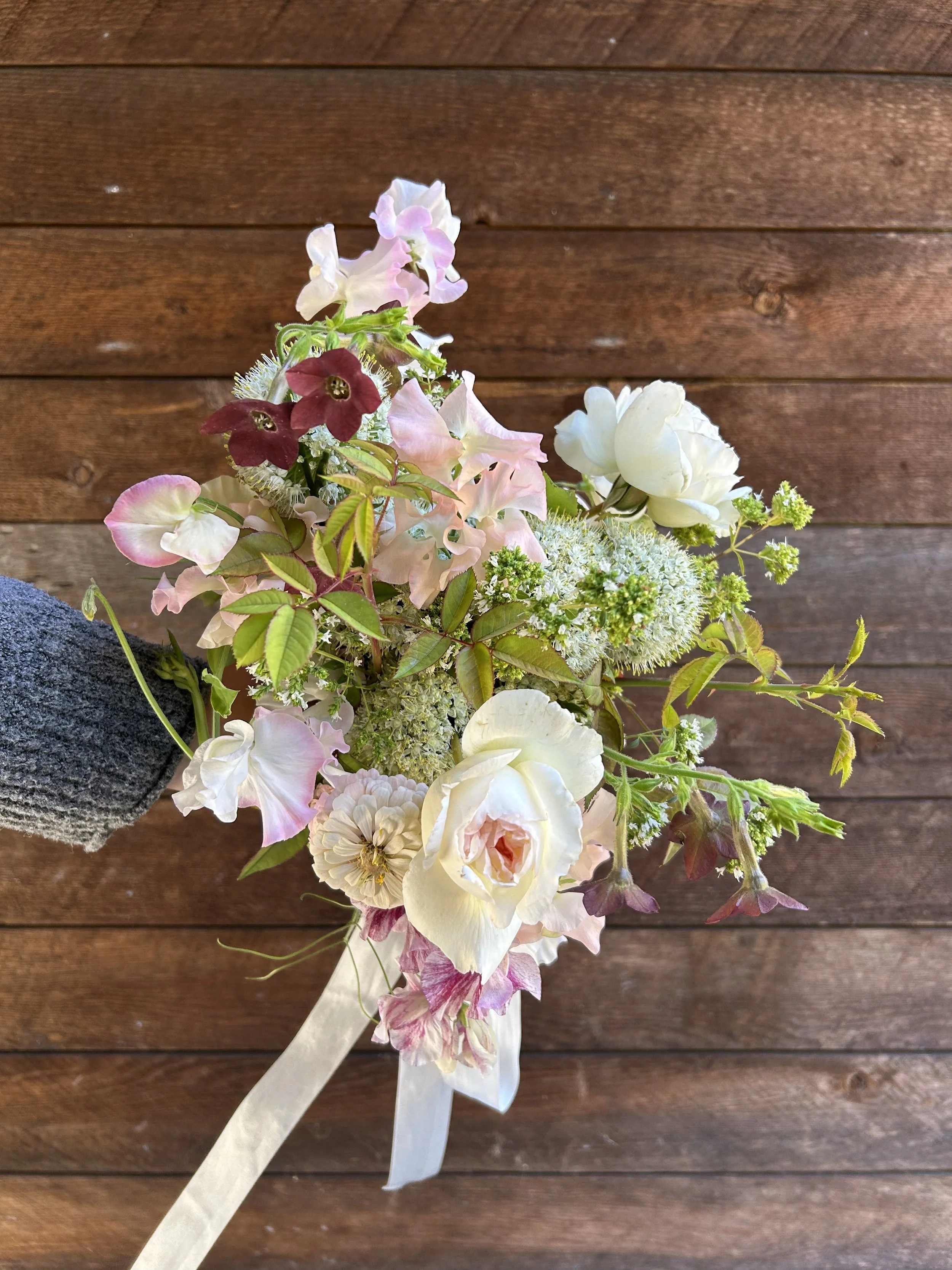 A bouquet of various pastel-colored flowers including roses, sweet peas, and other blooms, tied with a white ribbon, held by a person wearing a gray sweater, with a wooden background.