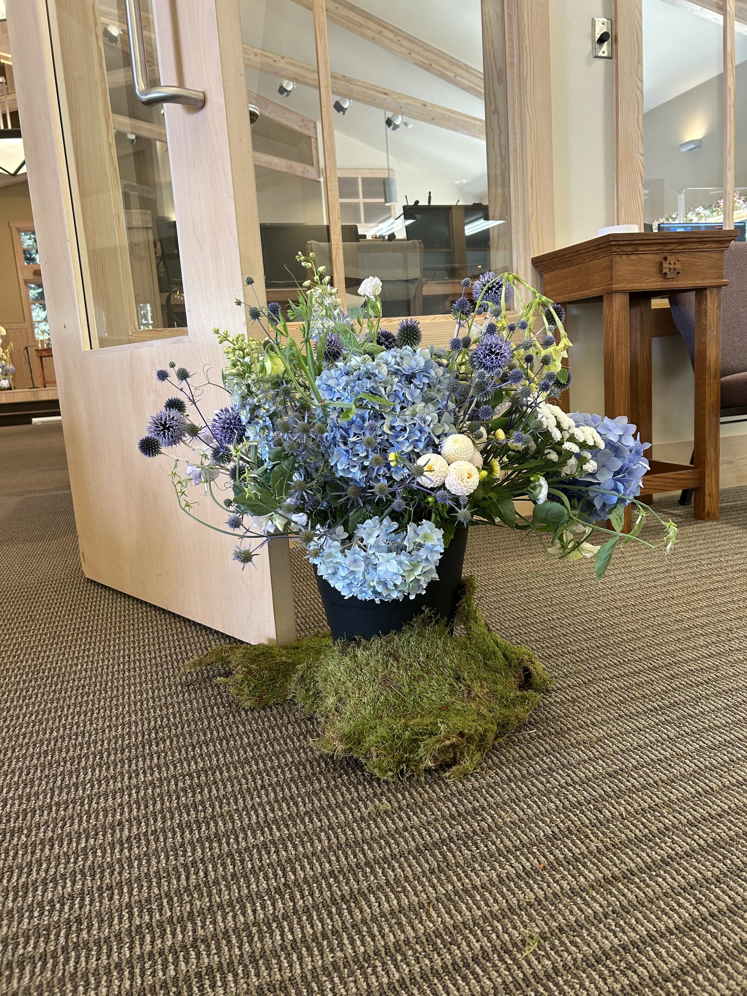 A floral arrangement of blue hydrangeas, white pom-pom flowers, purple thistles, and other greenery in a black pot, placed on a patch of moss on a carpeted floor inside a building.