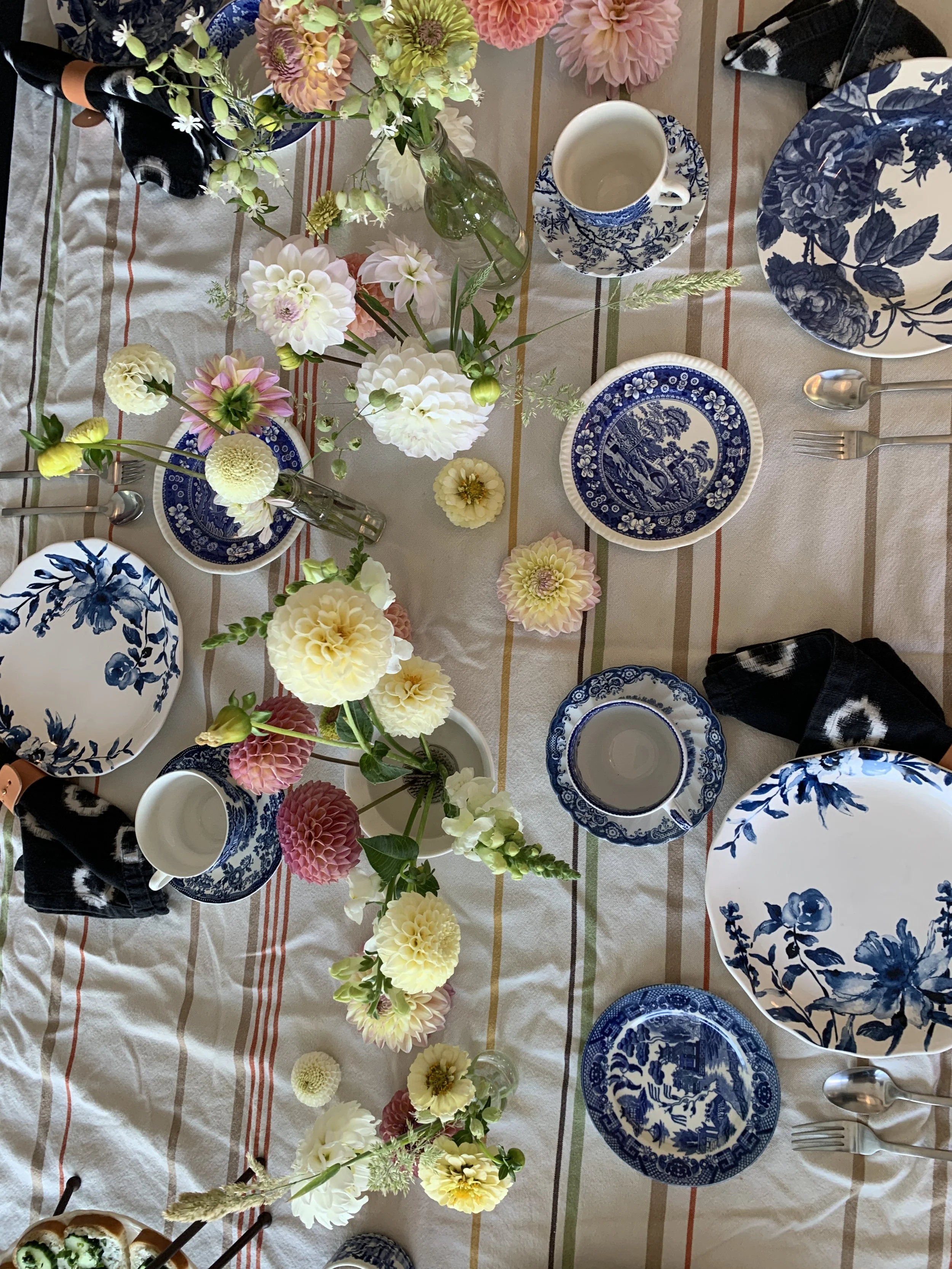 A table setting with floral arrangements, blue and white patterned dishes, cups, and silverware on a striped tablecloth.