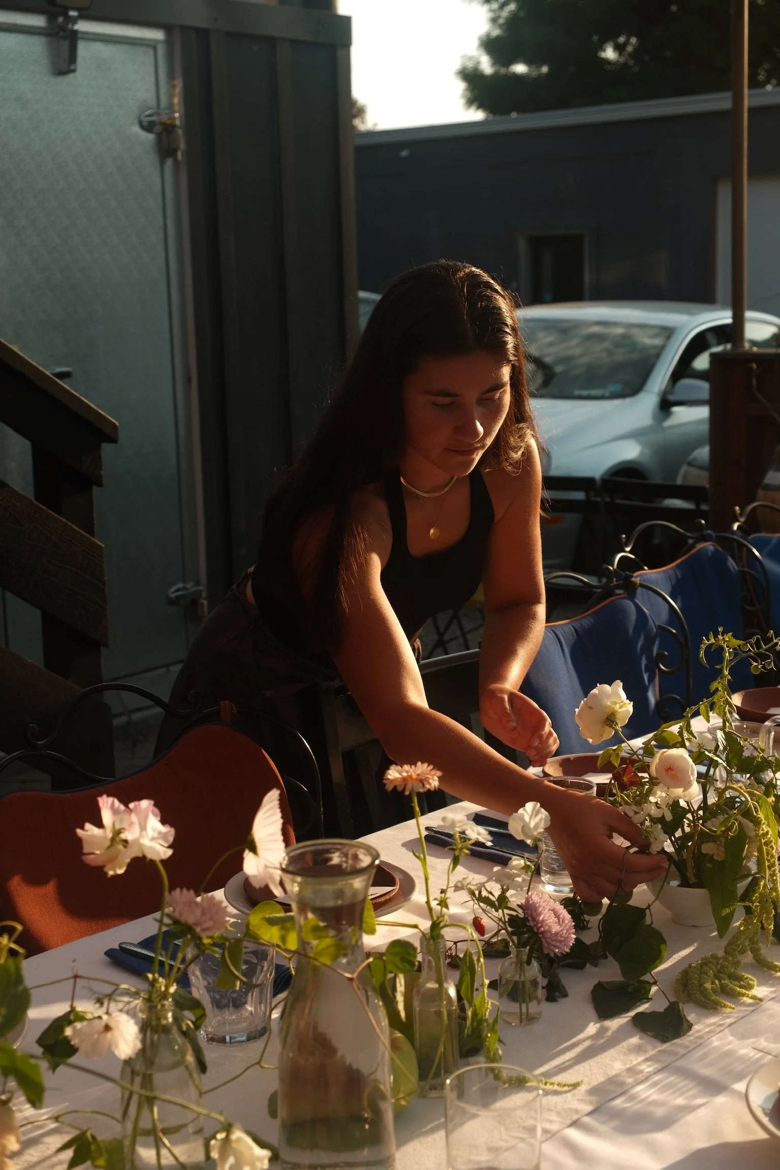 Woman arranging flowers on a table outdoors during late afternoon or sunset.