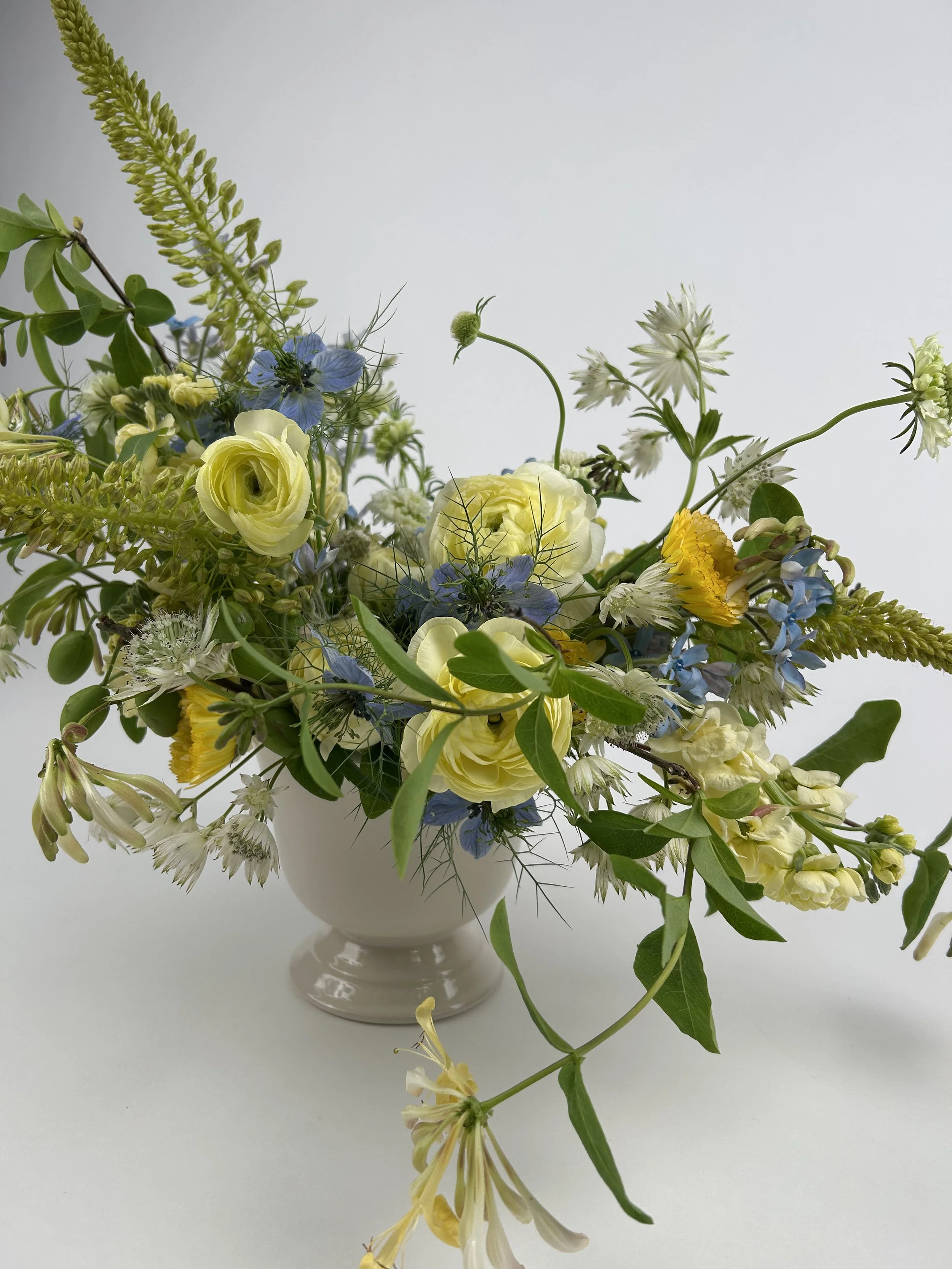 A bouquet of yellow, white, blue, and green flowers and foliage in a white vase set on a white surface against a plain white background.