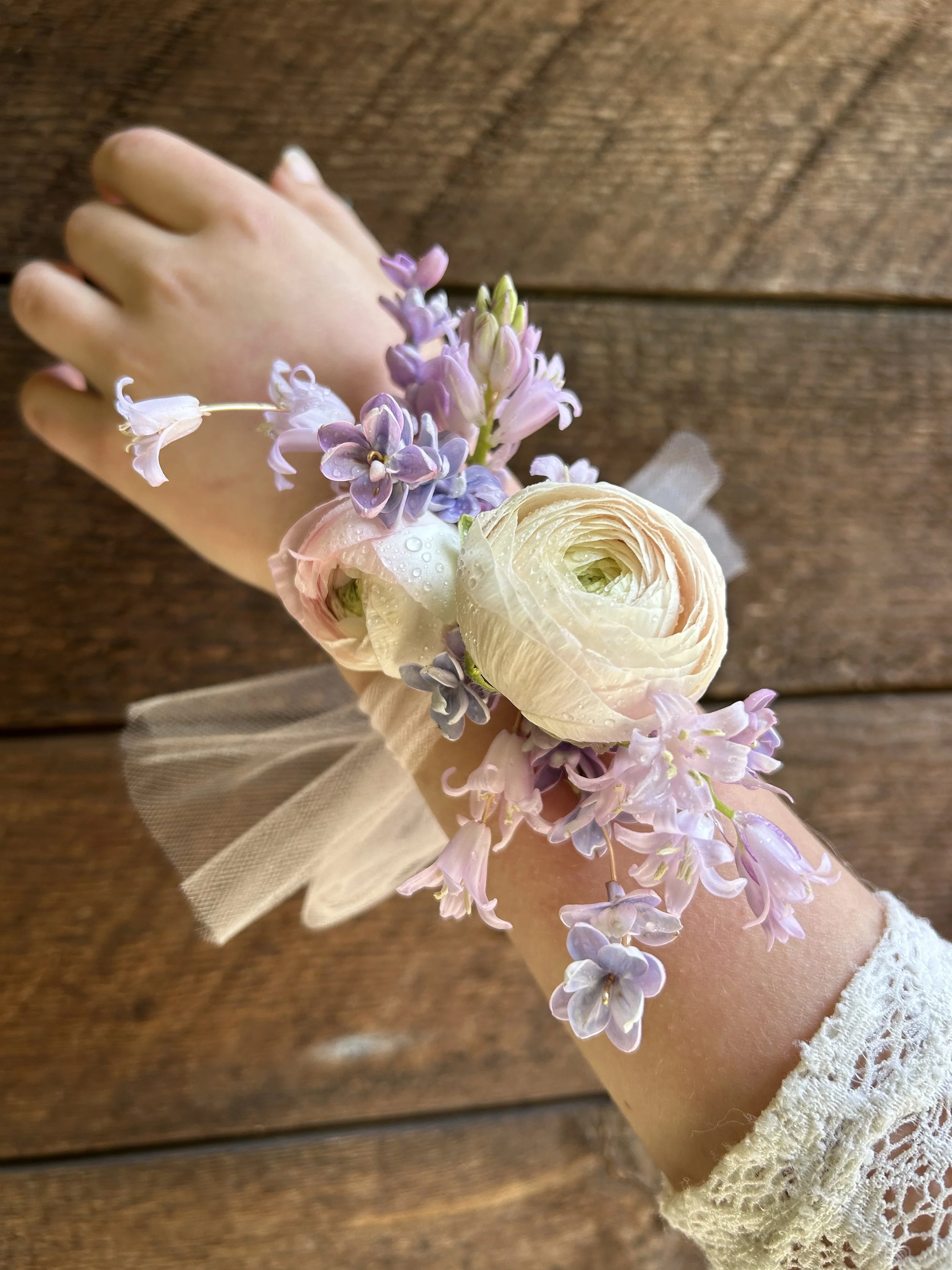 A person's forearm and hand wearing lace cuff, with a corsage of pink and purple flowers including ranunculus, hyacinth, and lilac, on a wooden surface background.