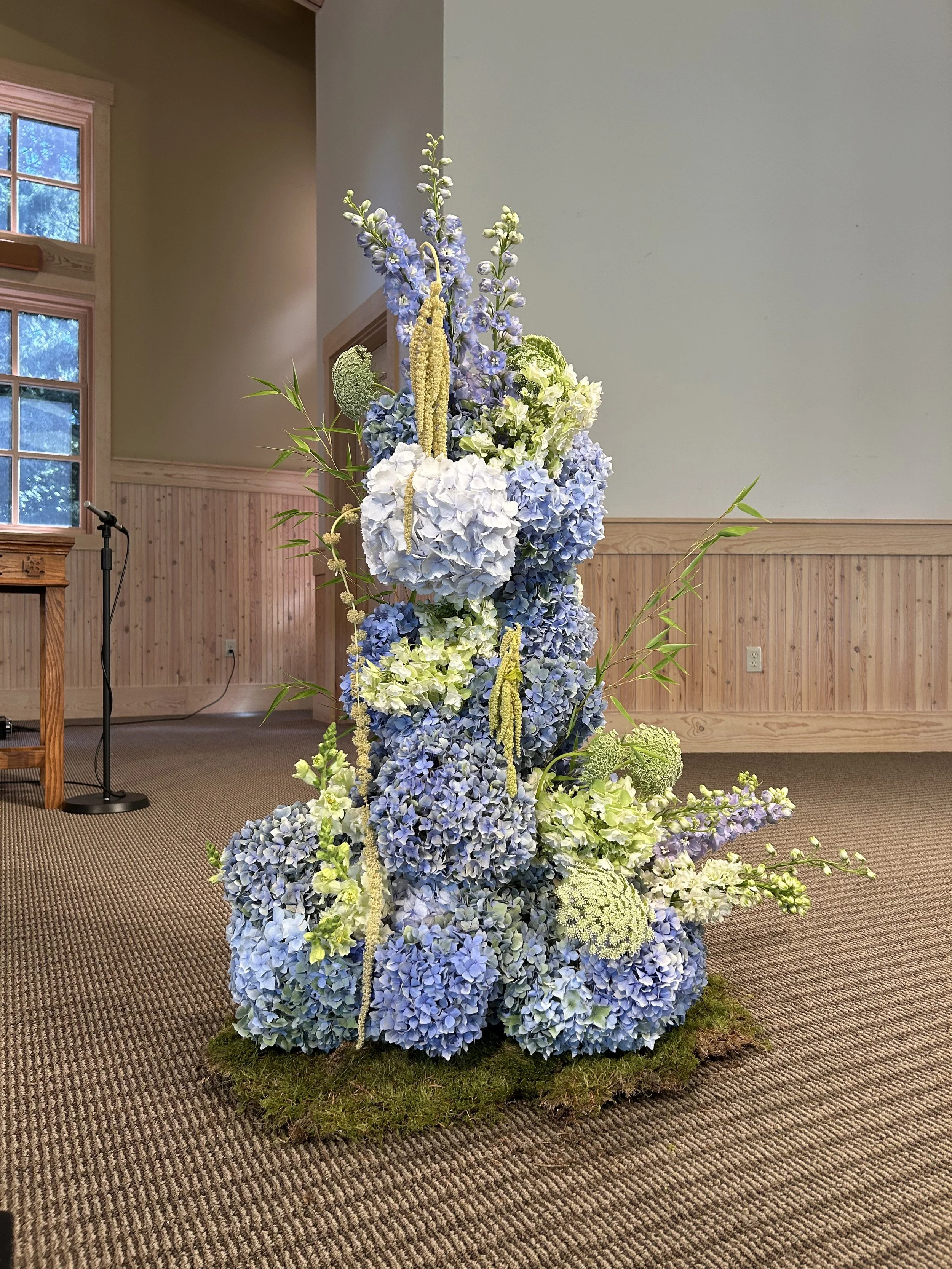 A floral arrangement featuring blue hydrangeas, white and green flowers, and tall greenery, placed on a mossy base in an indoor setting.