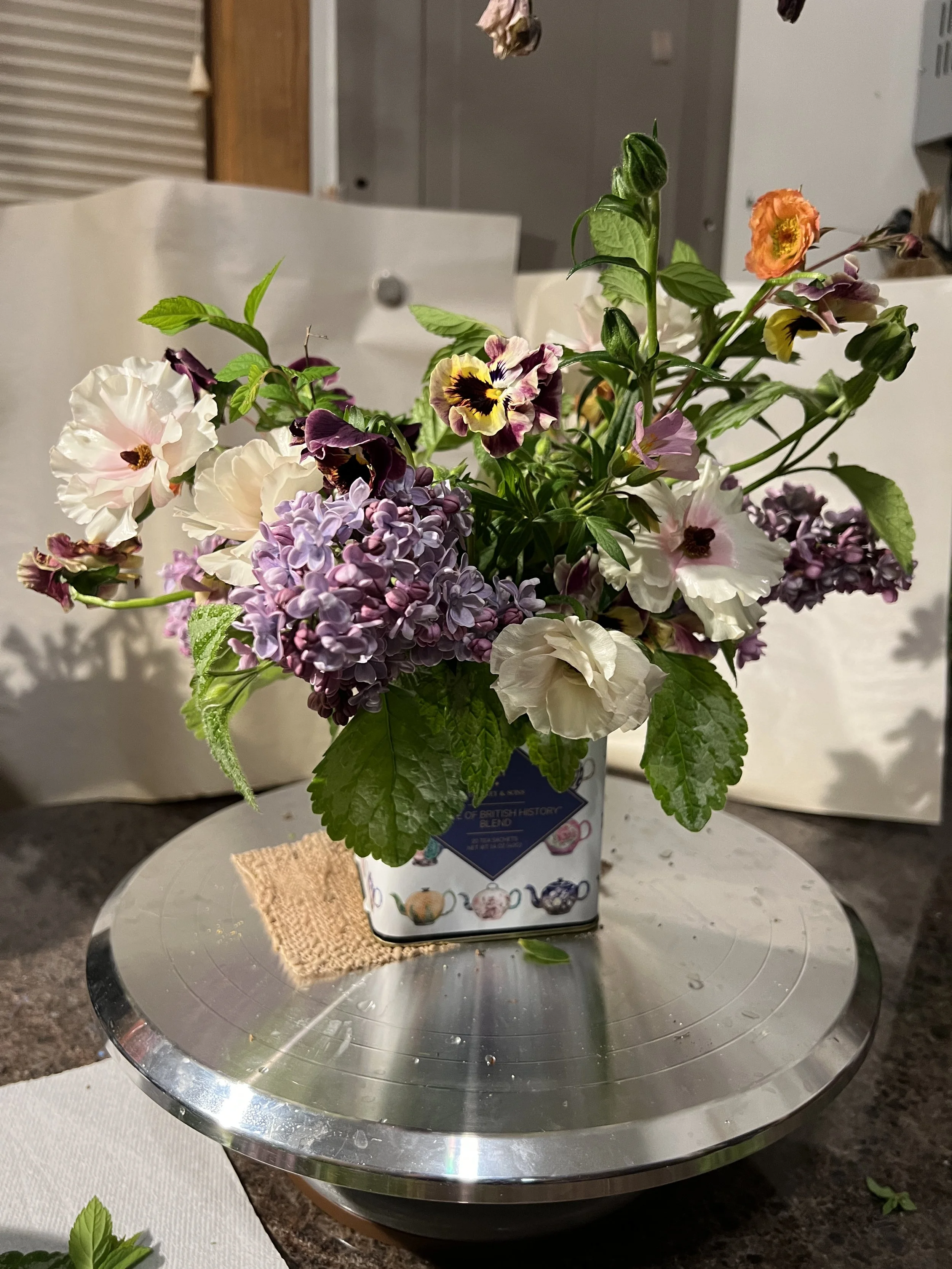 A bouquet of mixed flowers including violets, white and purple petunias, and orange blossoms arranged in a tea container.