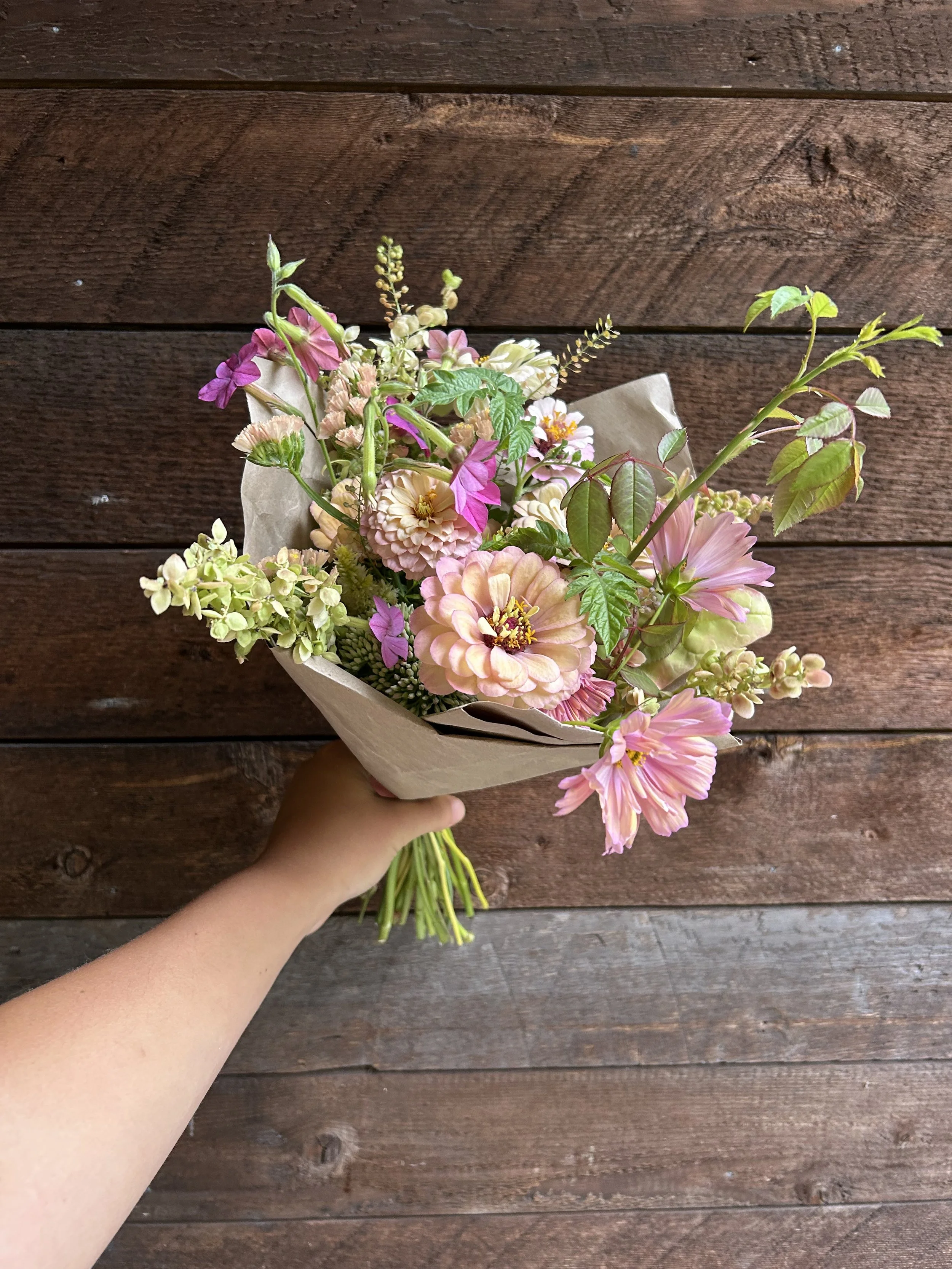 Hand holding a bouquet of pink and white flowers with green leaves, wrapped in brown paper, against a wooden background.