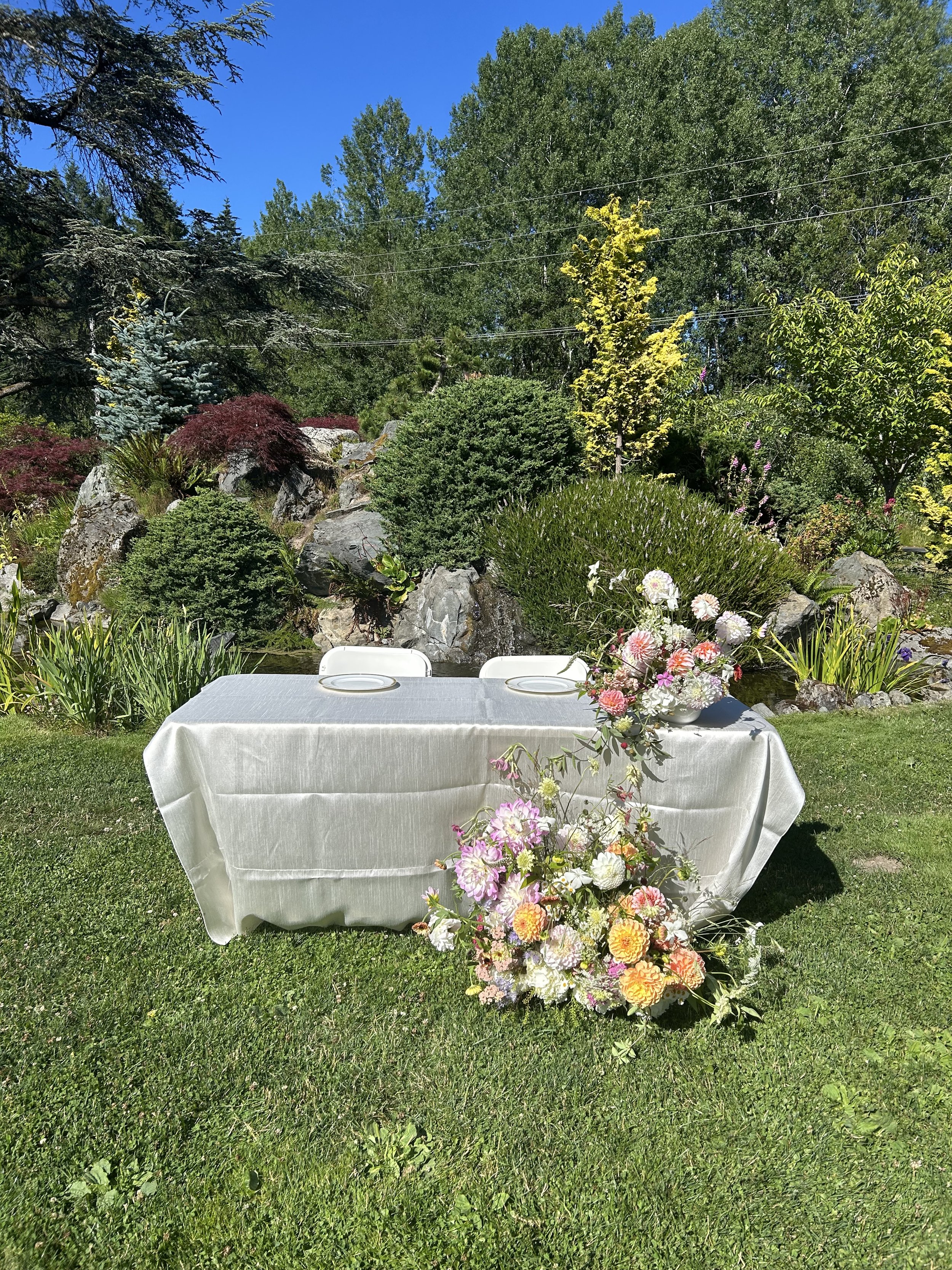 A long rectangular table covered with a white tablecloth, set outdoors on green grass. The table has two white chairs and a large floral arrangement with pink, white, and yellow flowers at one end. Behind the table is a garden with rocks and various 