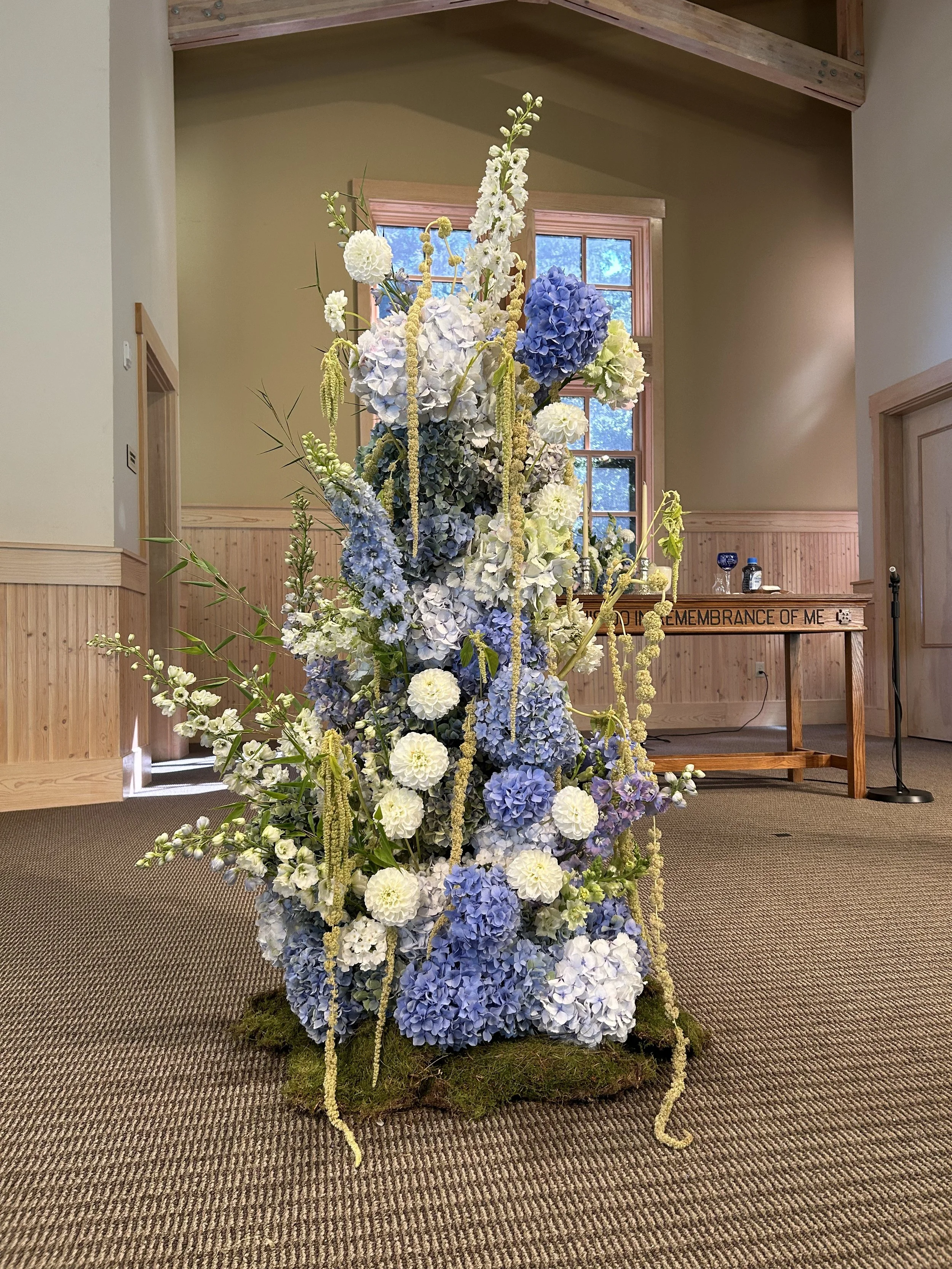 Tall floral arrangement of white and blue hydrangeas, white roses, and green foliage in a rustic indoor setting.