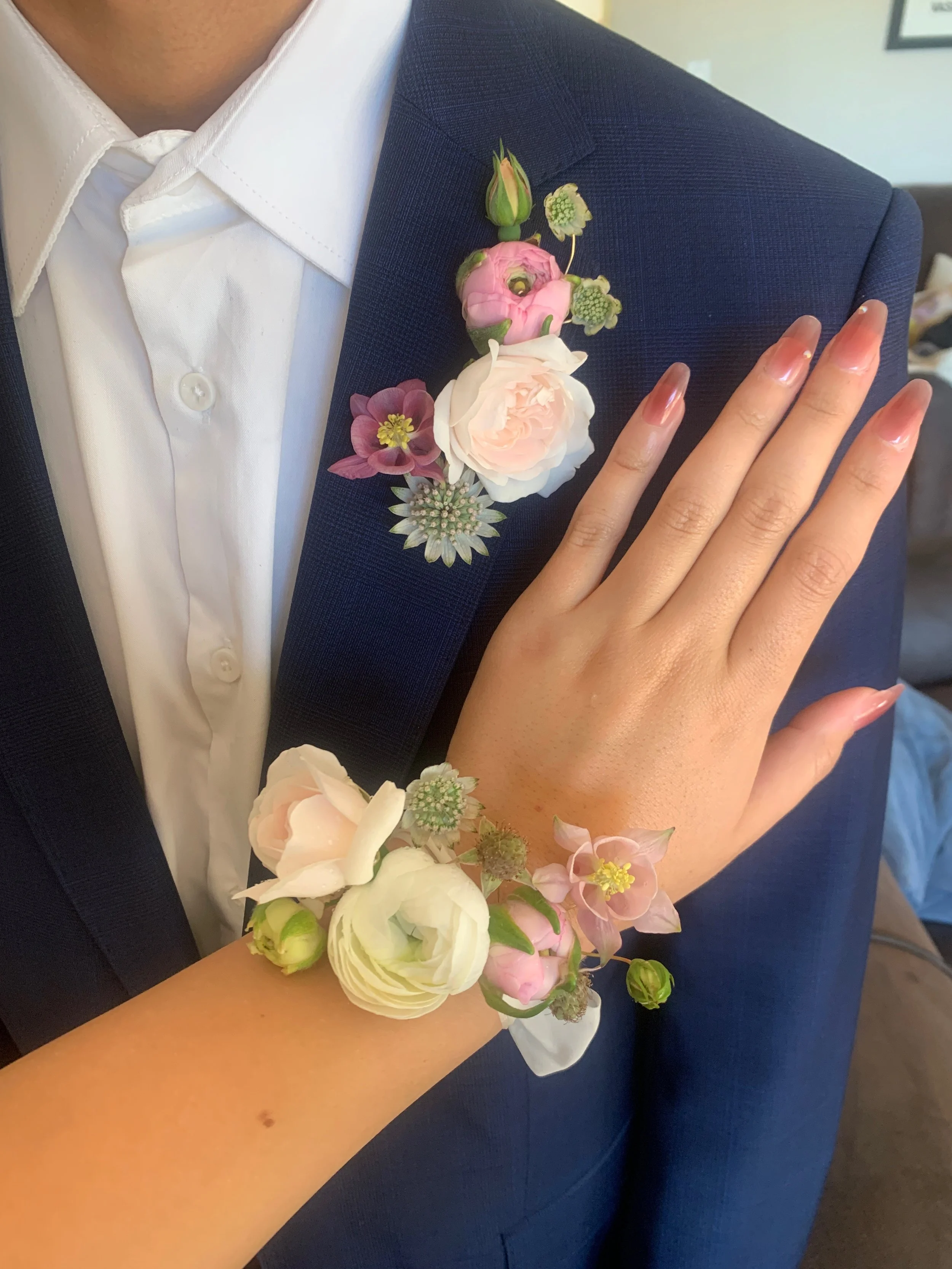A person wearing a dark blue suit and white shirt with a pink and white floral boutonniere on the lapel, also wearing a matching floral bracelet on the wrist, with a hand resting on the chest.