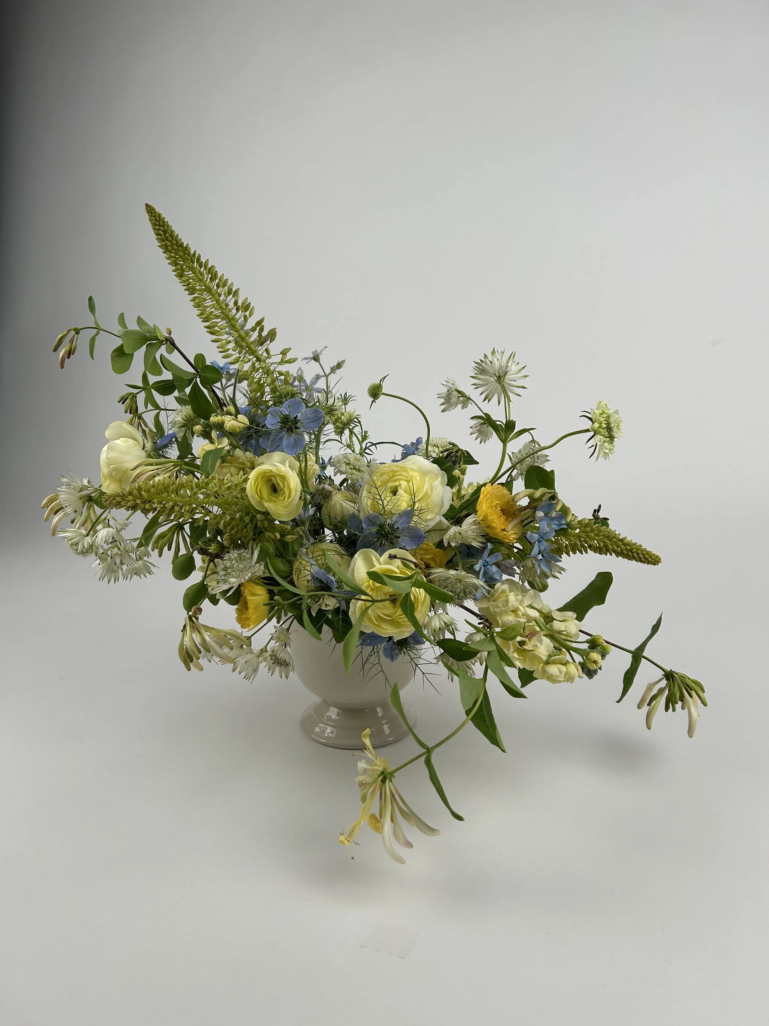 A white ceramic vase filled with a mixed floral arrangement of white, yellow, and blue flowers with green foliage on a white background.