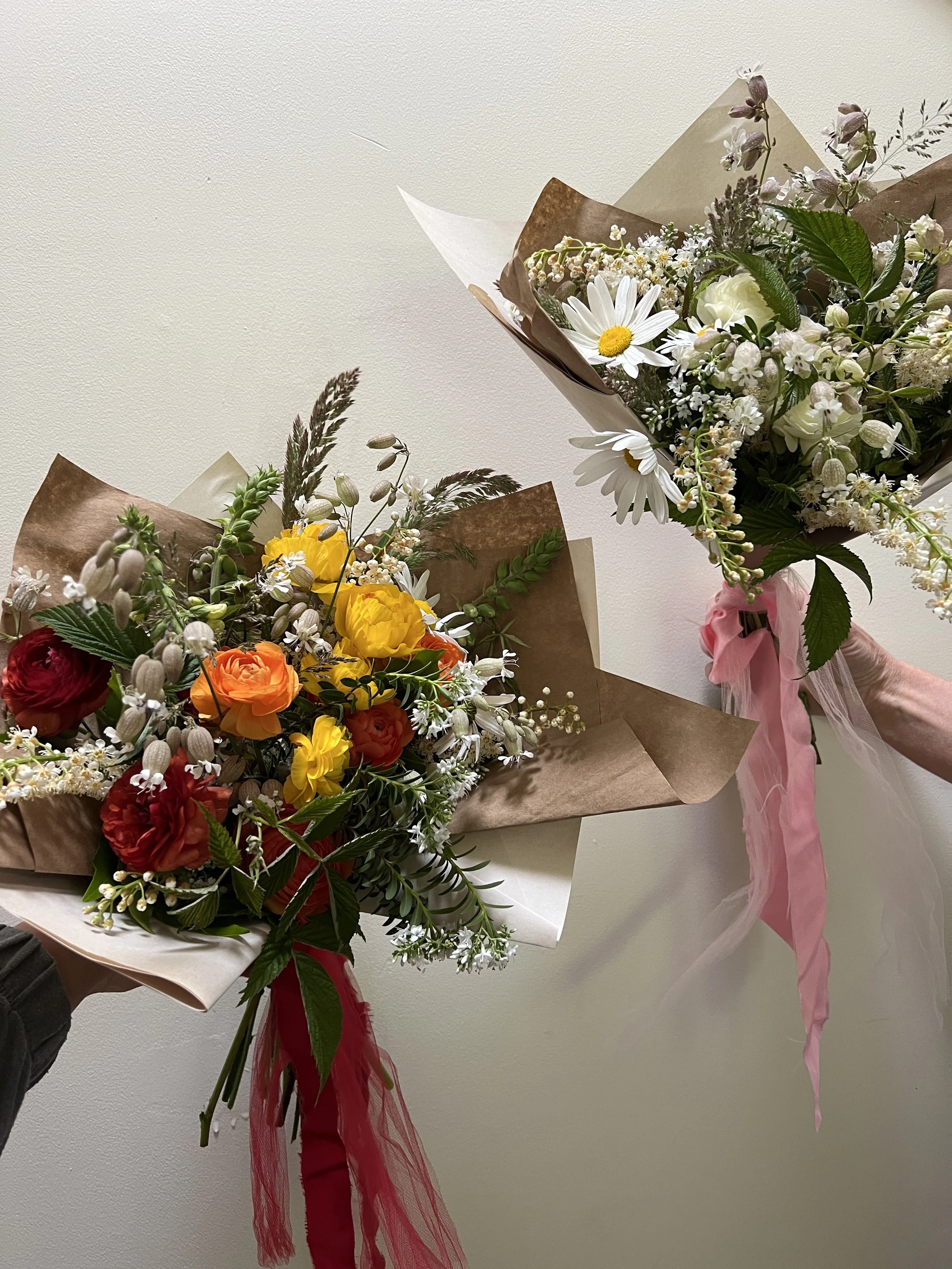 Two bouquets of colorful flowers, one with red, orange, and yellow blooms, and the other with white daisies, baby's breath, and greenery, both wrapped in brown wrapping paper with pink ribbons.