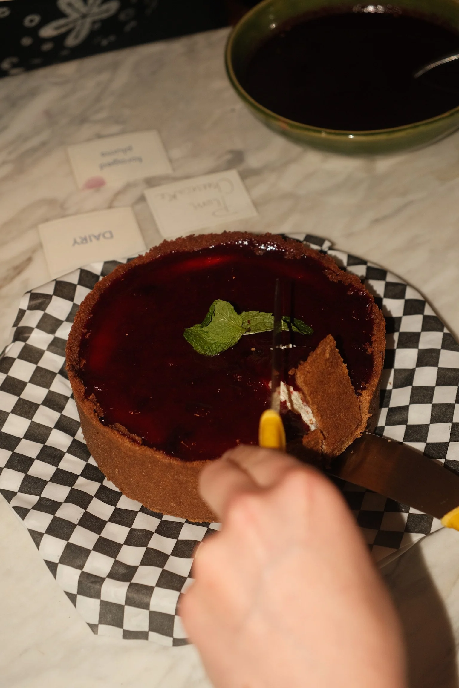 A person cutting into a round chocolate tart with a mint leaf garnish, placed on black and white checkered paper, with a bowl of dipping sauce in the background.