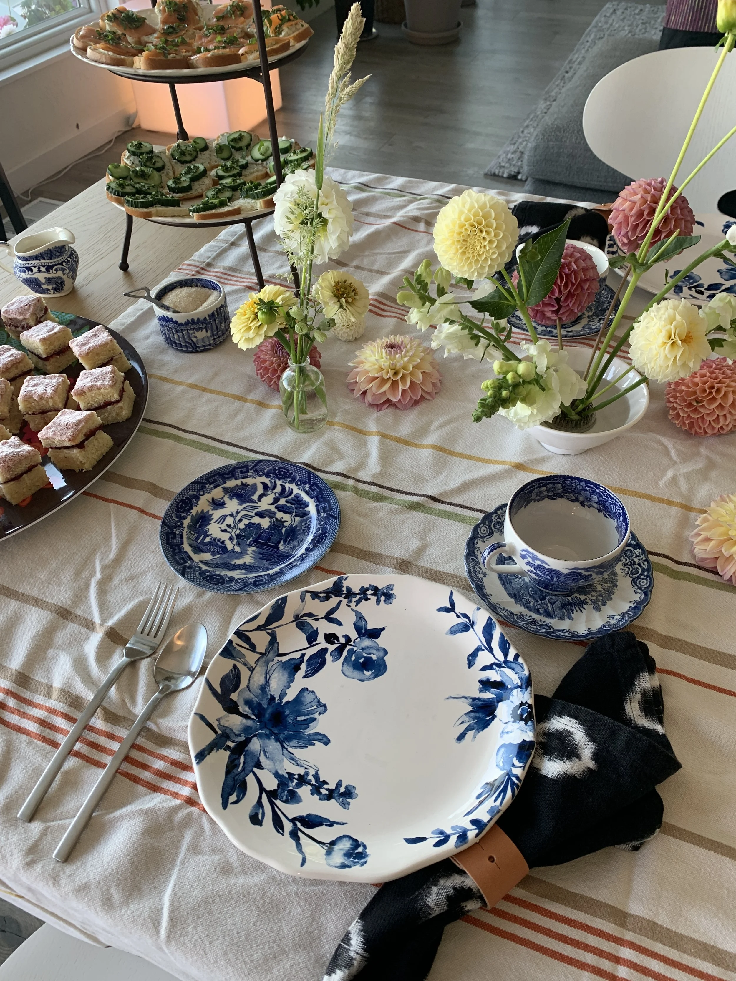 A table set with blue and white floral patterned dishes, a black cloth napkin, a fork, knife, and spoon, a teacup with saucer, and decorated with pink and white flowers. There are also two-tiered stands with finger foods and a variety of desserts.