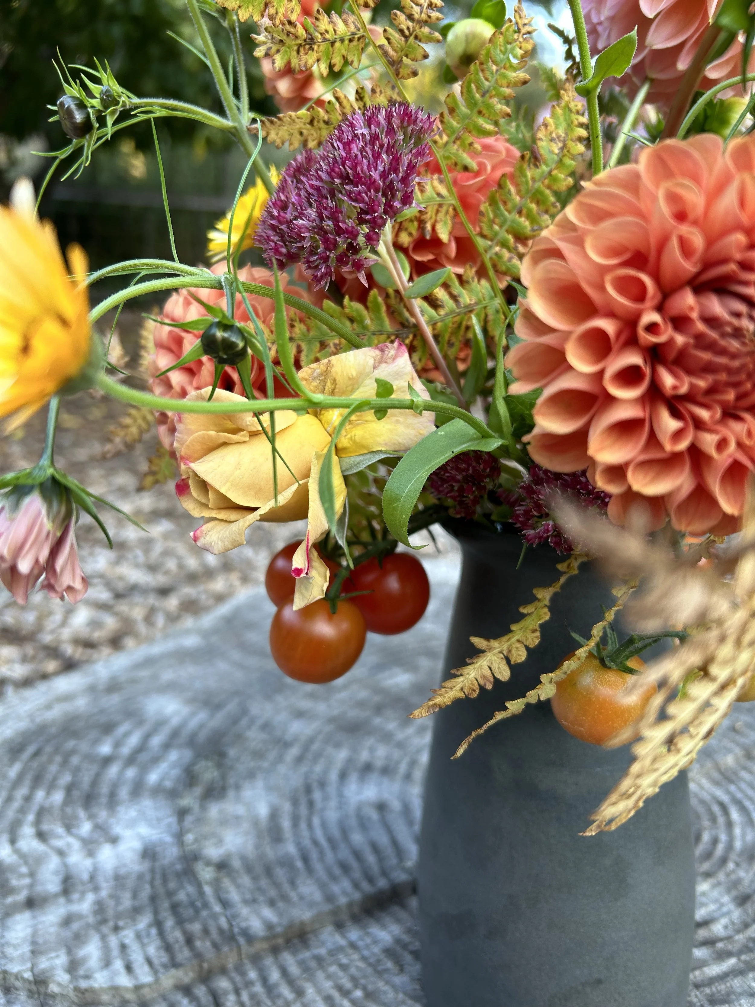 A close-up of a colorful flower bouquet in a black vase on a wooden surface, including pink and orange flowers, small tomatoes, and green foliage.