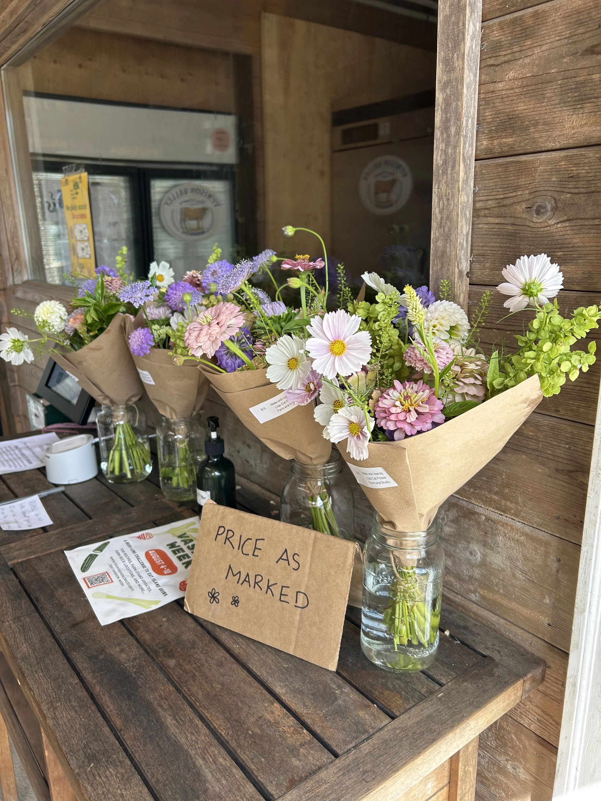 Three small bouquets of mixed pink, purple, and white flowers in glass jars on a wooden table outside a rustic building. A cardboard sign reads 'Price as marked'.