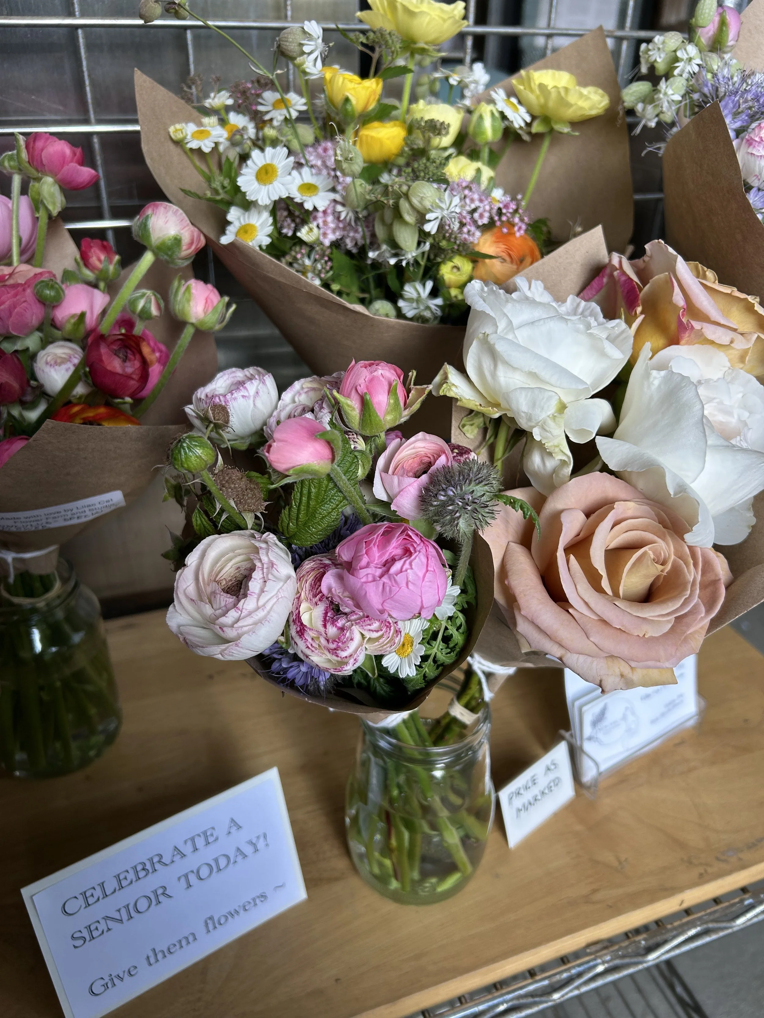 Assorted flower bouquets in glass jar on wooden table, with signs reading 'Celebrate a senior today!' and 'Prices to maker?'.