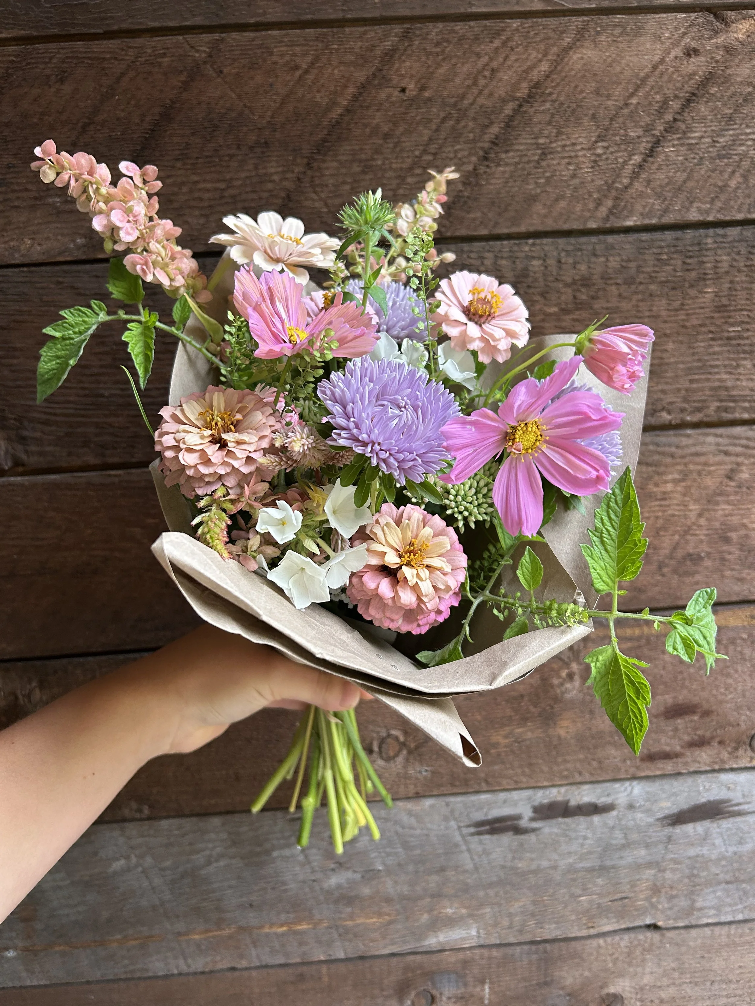 Hand holding a bouquet of pink, purple, and white flowers wrapped in beige paper, against a wooden background.