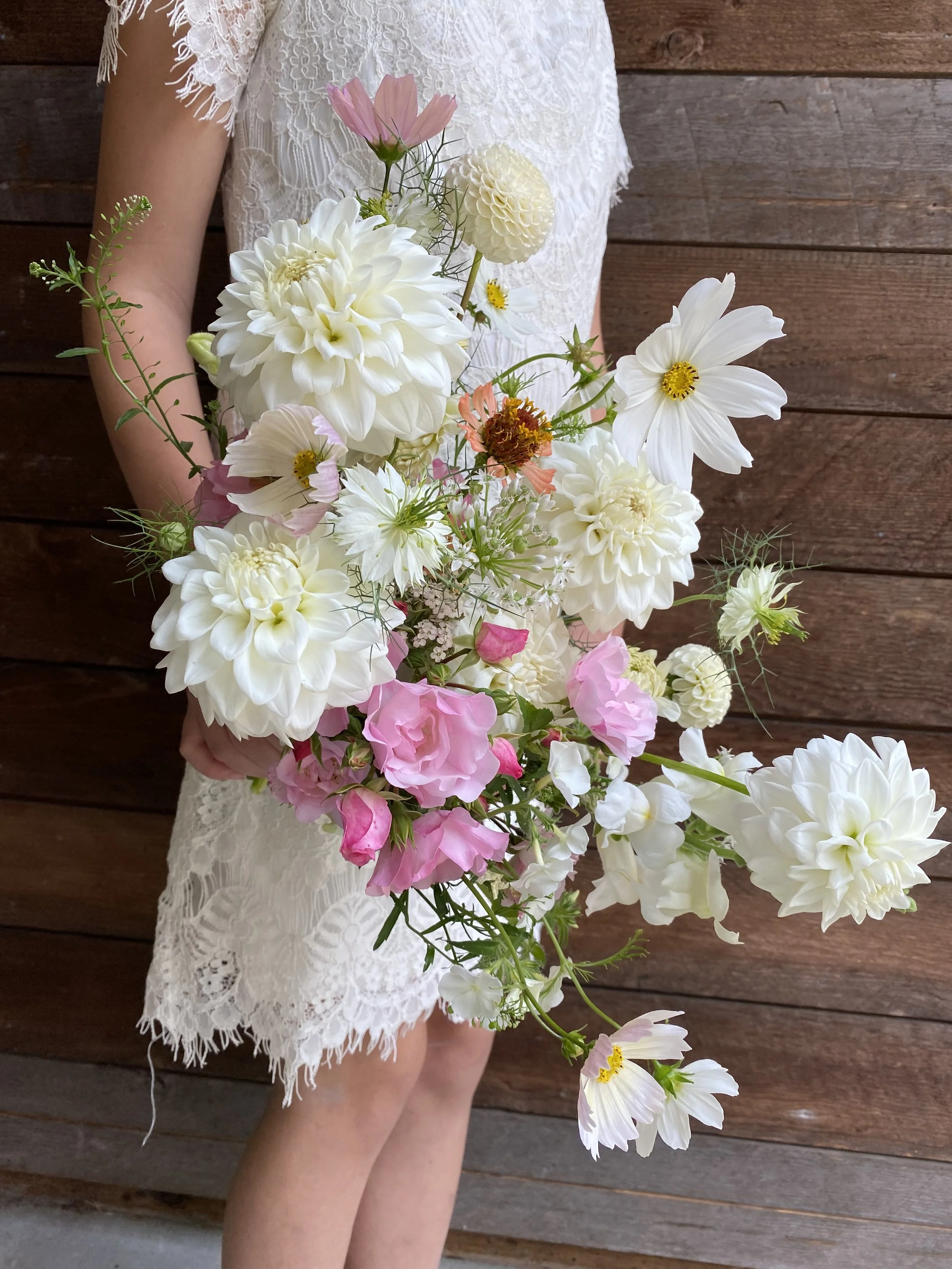 Person in a white lace dress holding a large bridal bouquet of white, pink, and peach flowers against a wooden background.