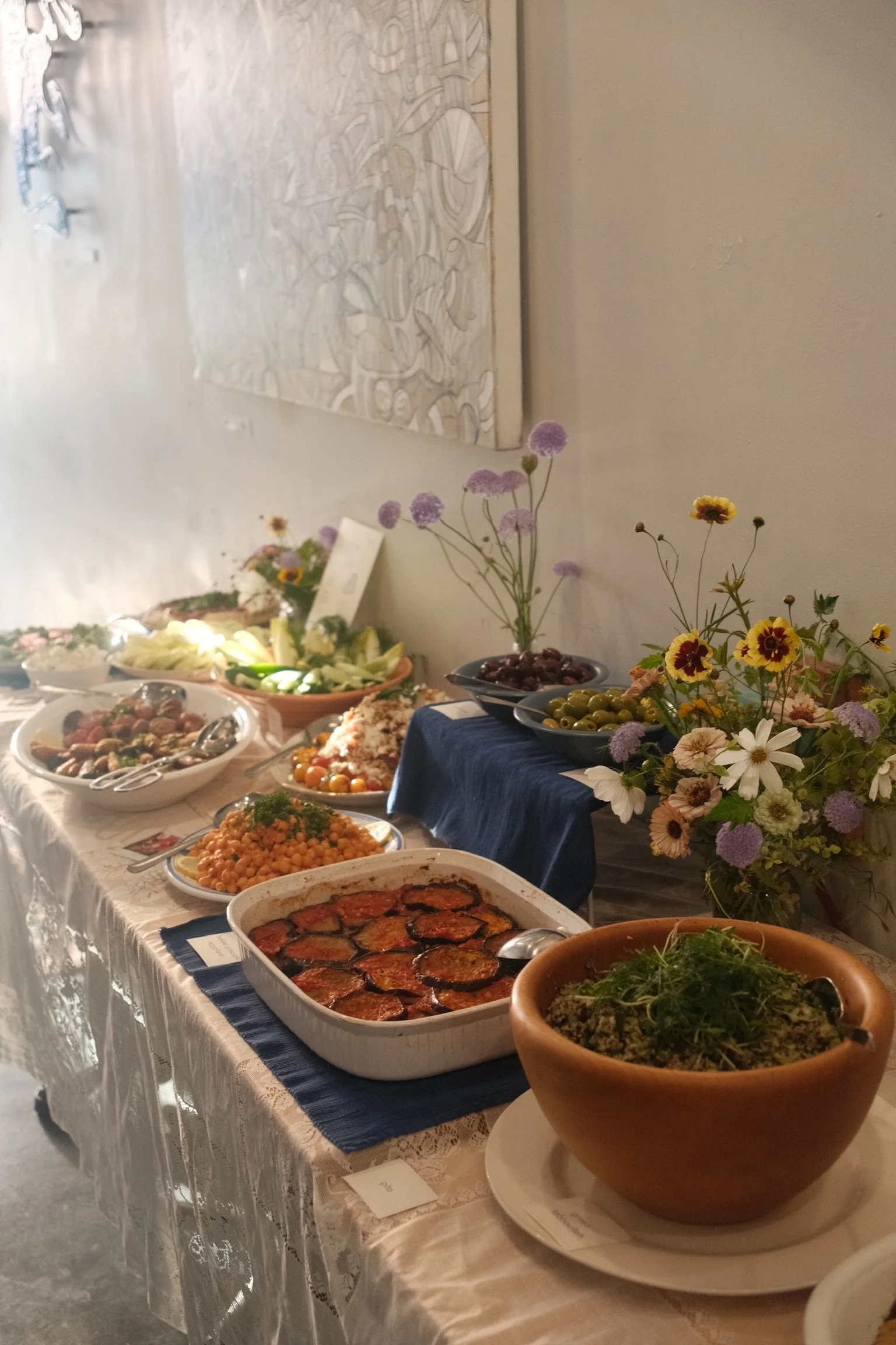 A buffet table with various dishes, including salads, a casserole with roasted vegetables, and a large bowl of greens. The table is decorated with colorful flowers in vases and has a lace tablecloth.