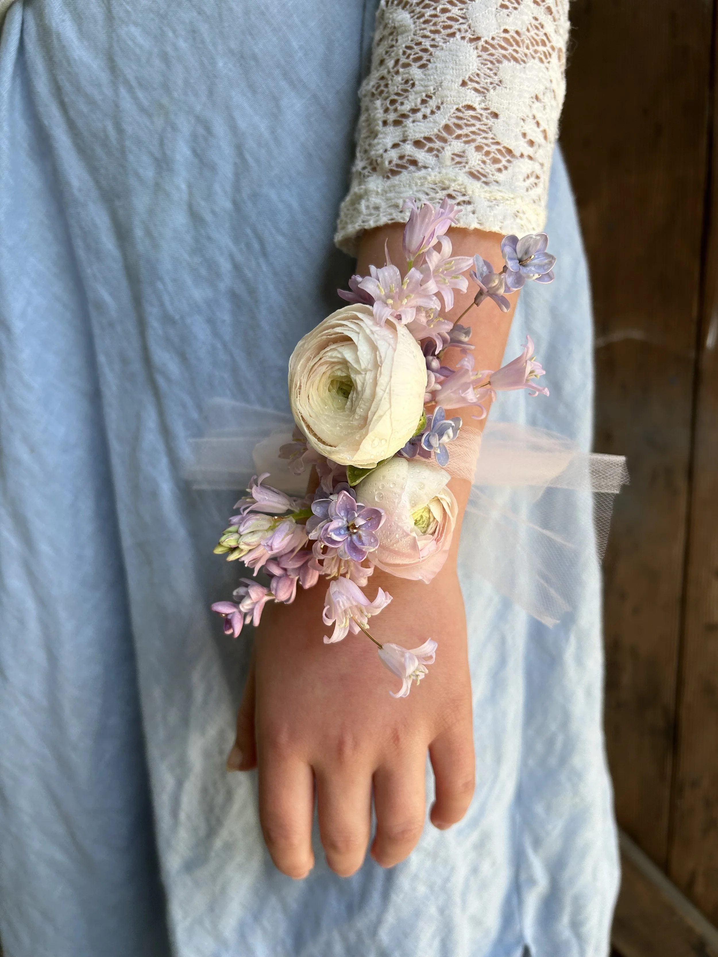 Person wearing a lace sleeve dress with a wrist corsage of white and purple flowers and tulle ribbon.