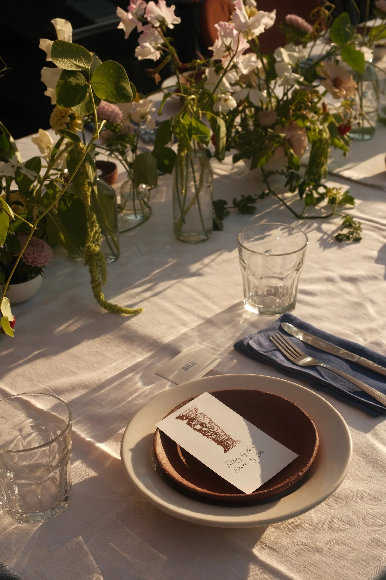 A table setting with a white tablecloth, glassware, a blue cloth napkin with a fork and knife, a plate with two brown pottery pieces, and a card with an illustration and text. The background features floral arrangements in glass vases with pink and p