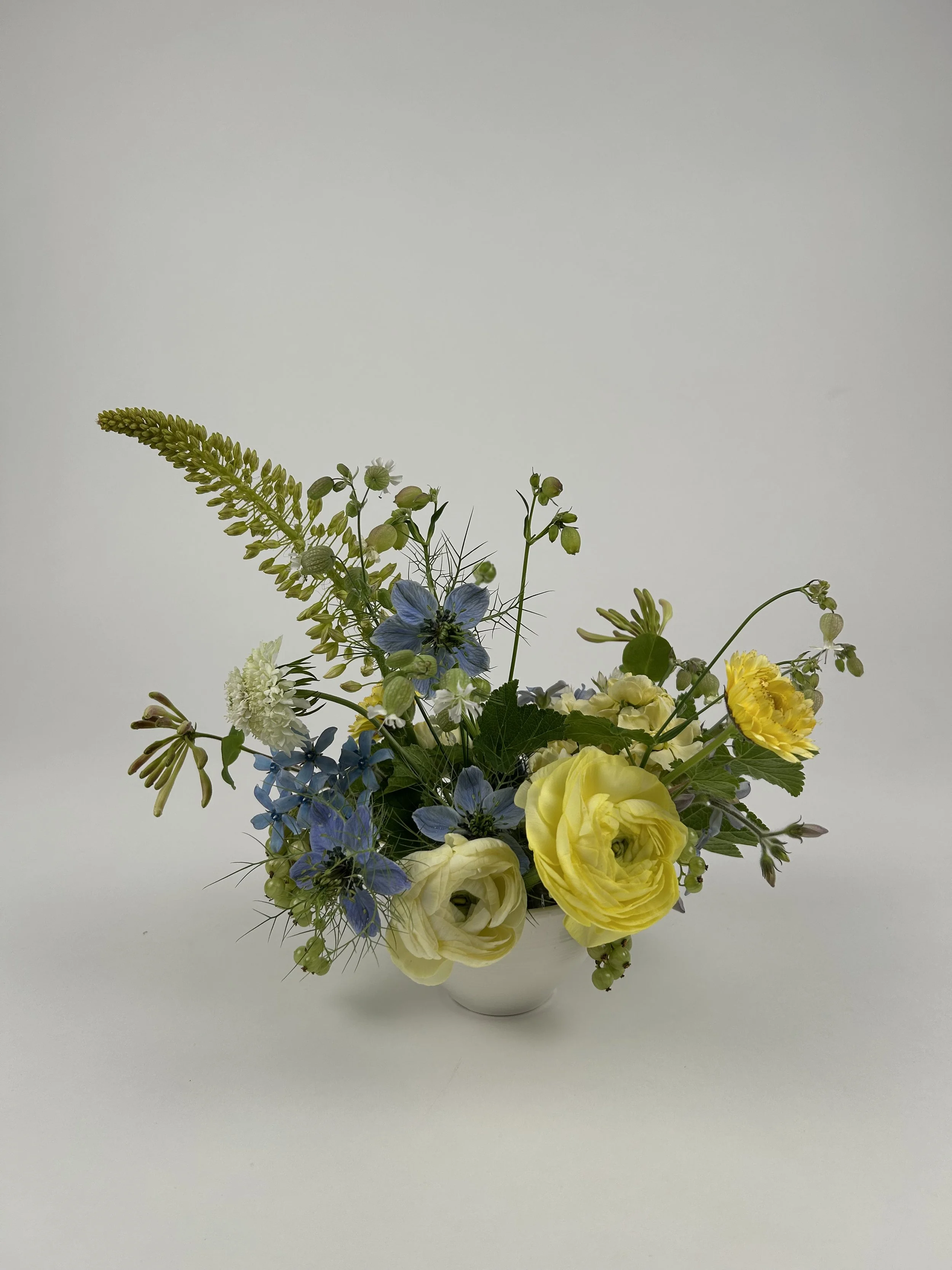 A floral arrangement with yellow and blue flowers in a white vase on a plain white background.