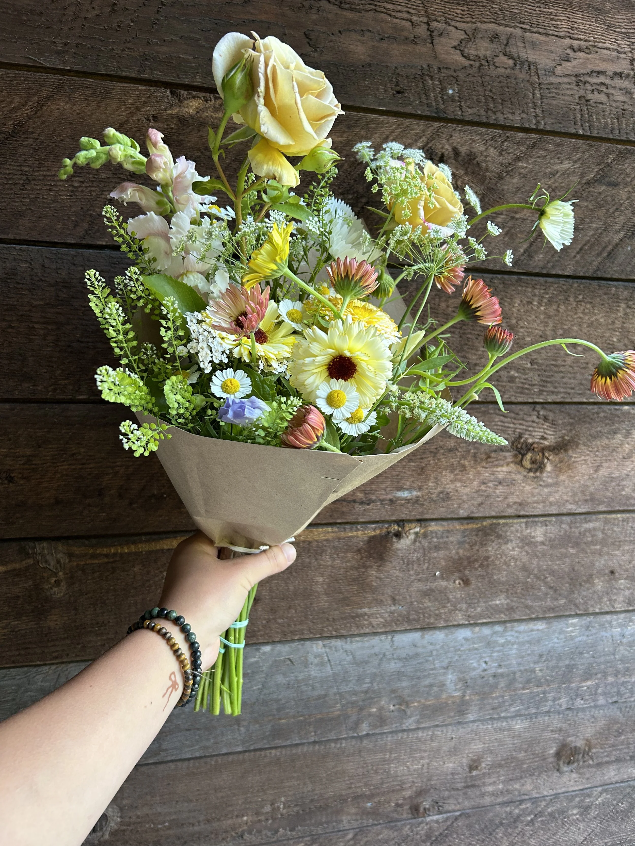 A hand holding a bouquet of various colorful flowers including yellow roses, daisies, and other mixed flowers in front of a wooden wall.