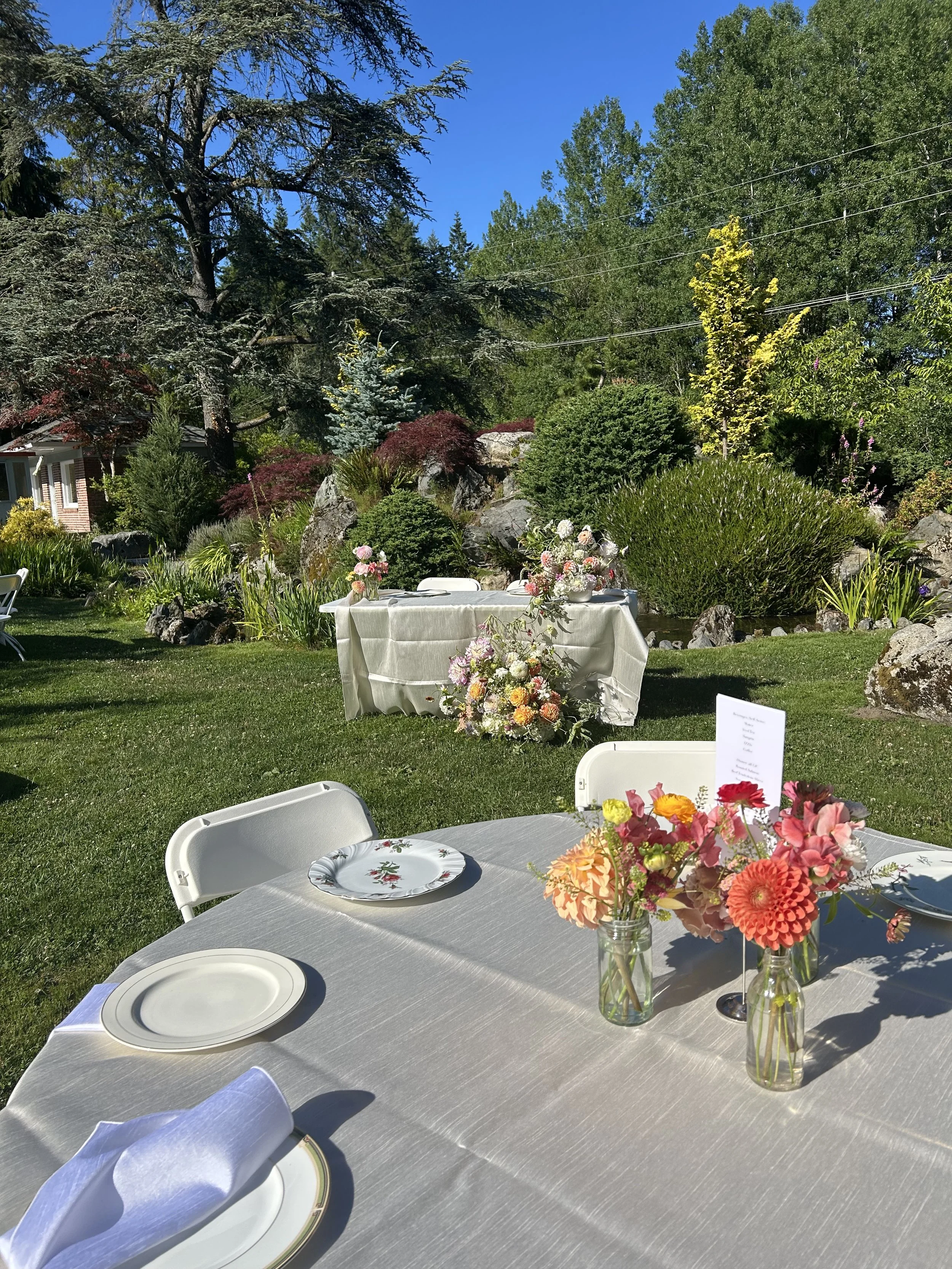 Outdoor garden setup with tables decorated with flowers, set for a celebration or event, surrounded by trees and greenery under a clear blue sky.