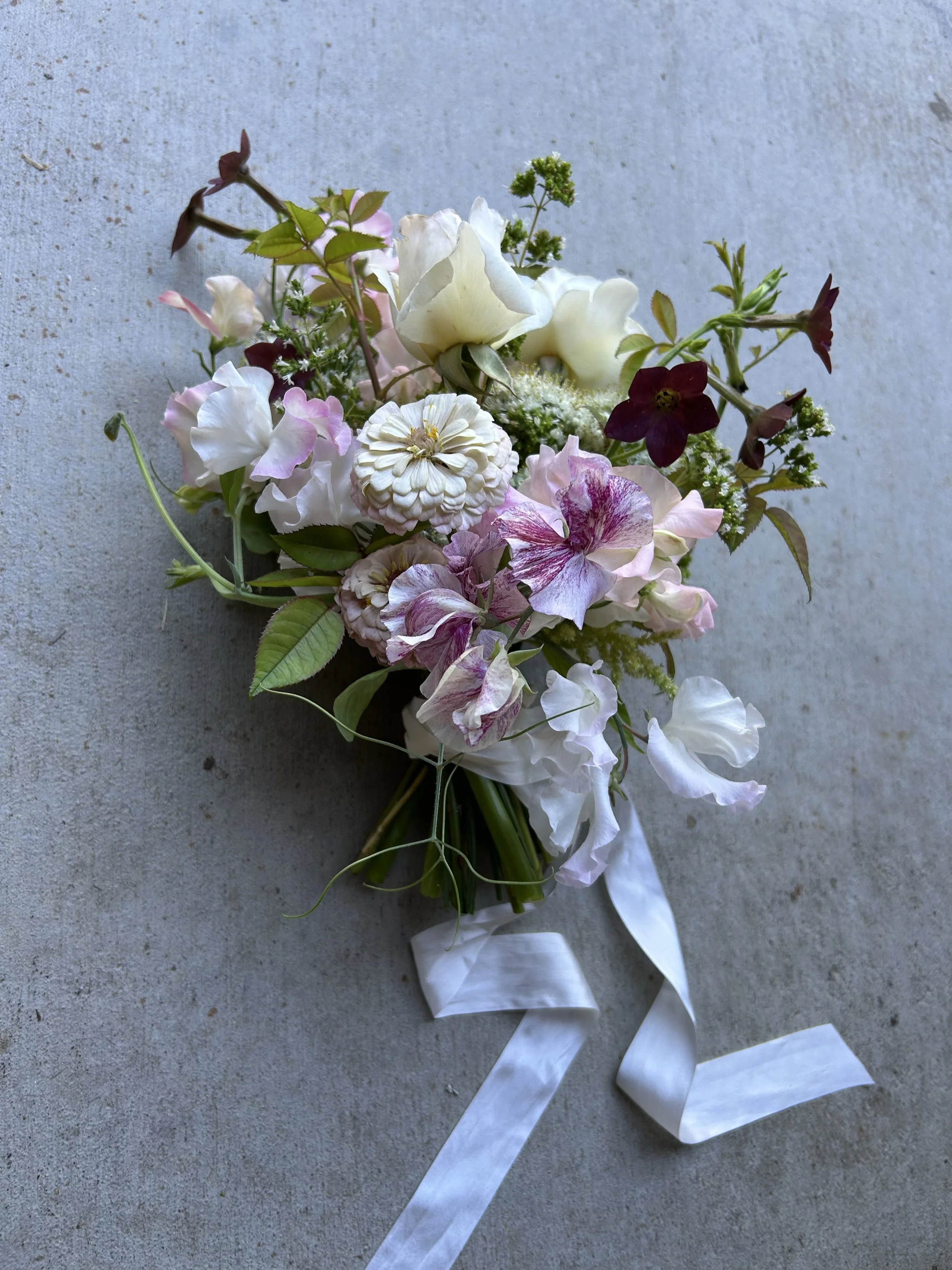 A bouquet of assorted pink, white, and purple flowers with green leaves and a white ribbon on a gray surface.