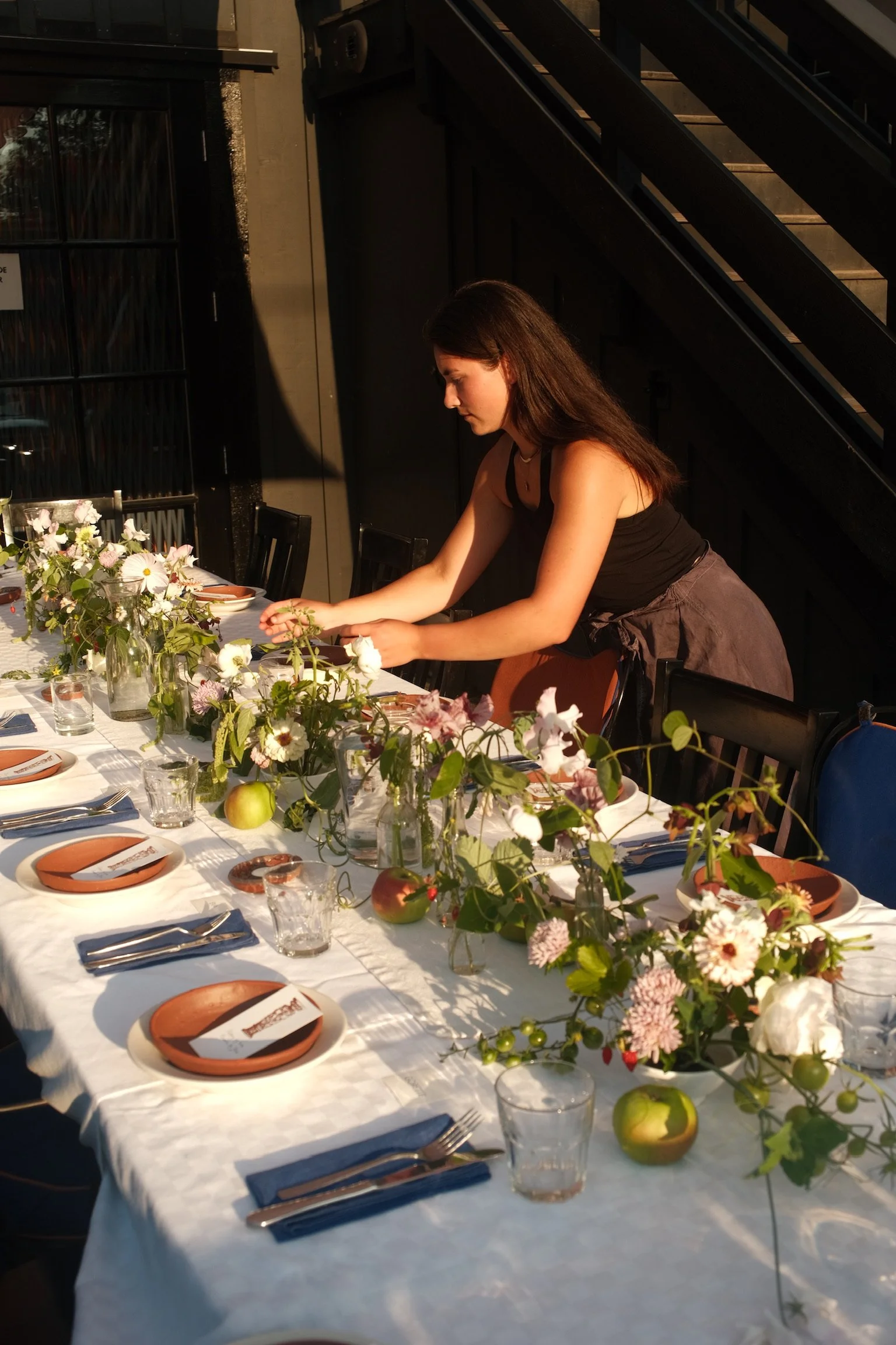 A woman preparing a decorated table with flowers, apples, and tableware for a meal or event.