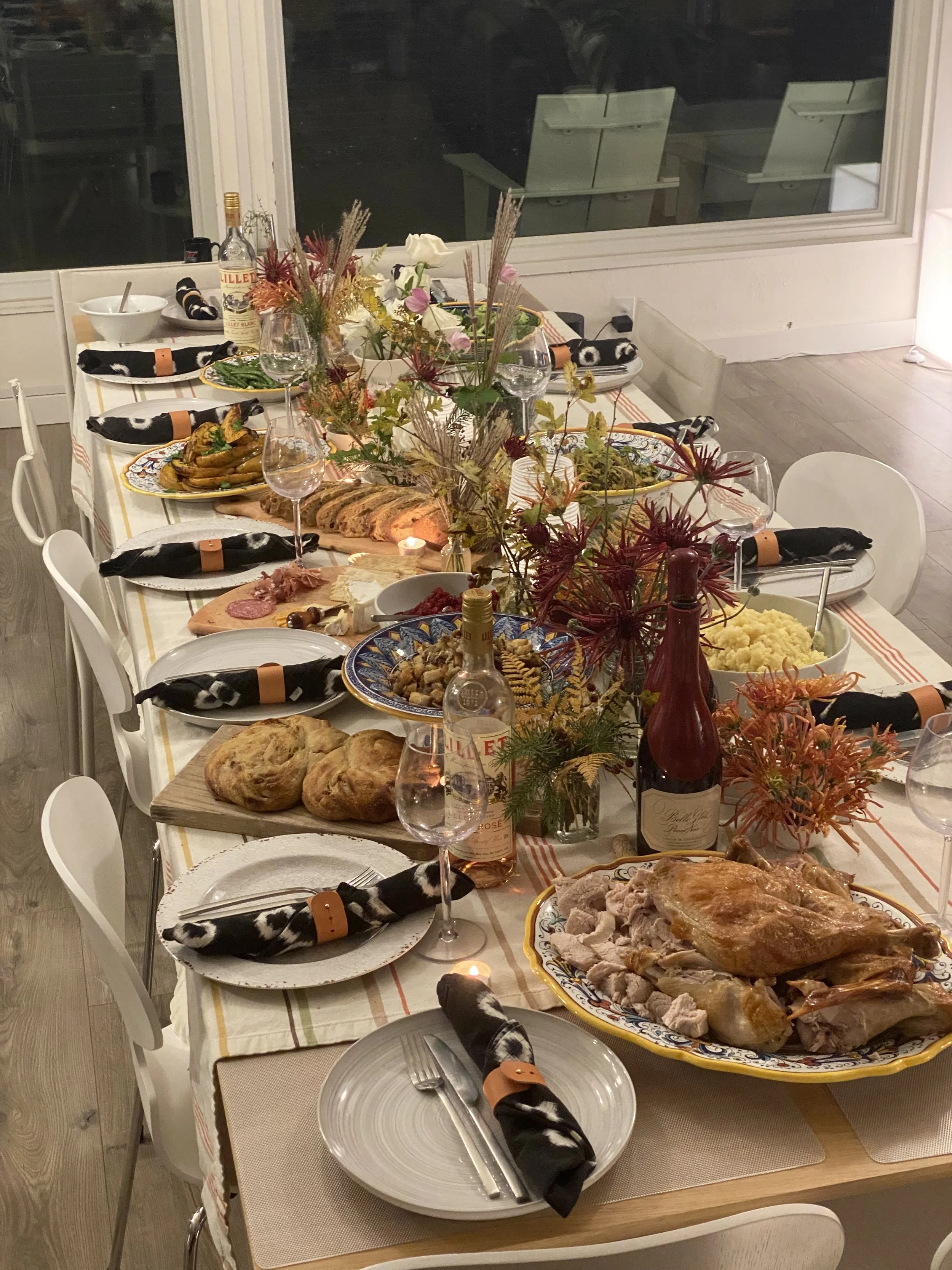 A festive dinner table set for a special occasion, featuring a roasted turkey, various side dishes, wine bottles, glasses, candles, and a floral centerpiece, with black and white napkins at each place setting.