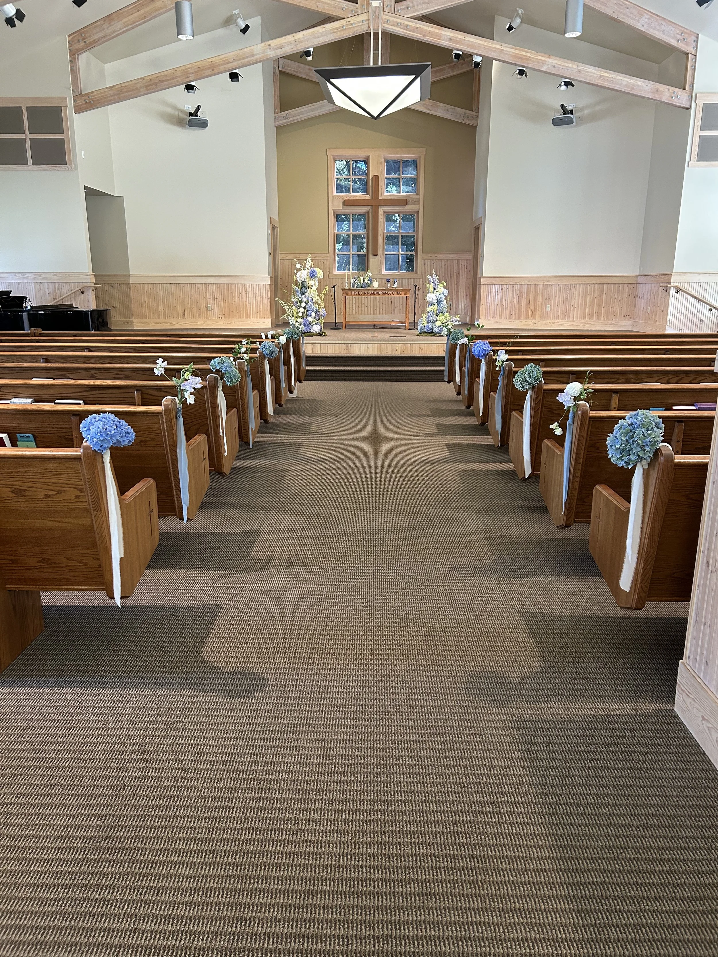 An indoor wedding chapel with wooden pews decorated with blue and white flowers, leading to an altar with a cross, floral arrangements, and a window behind it.