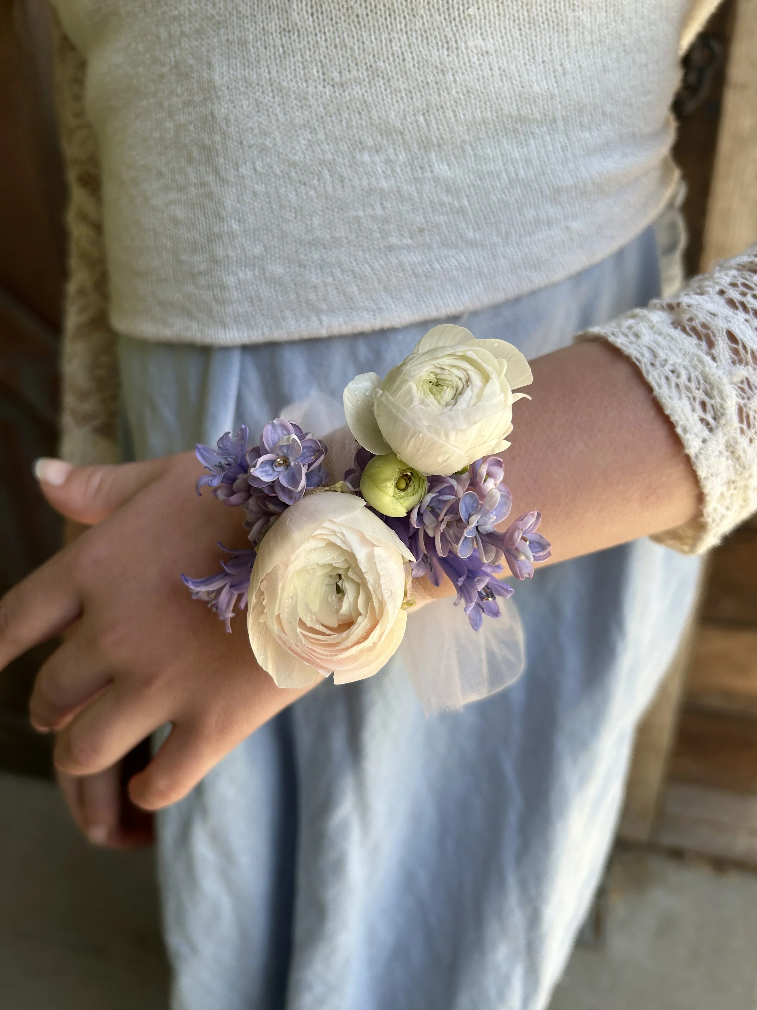 A person wearing a lace bracelet with a floral corsage featuring white ranunculus and purple hyacinths on their wrist.
