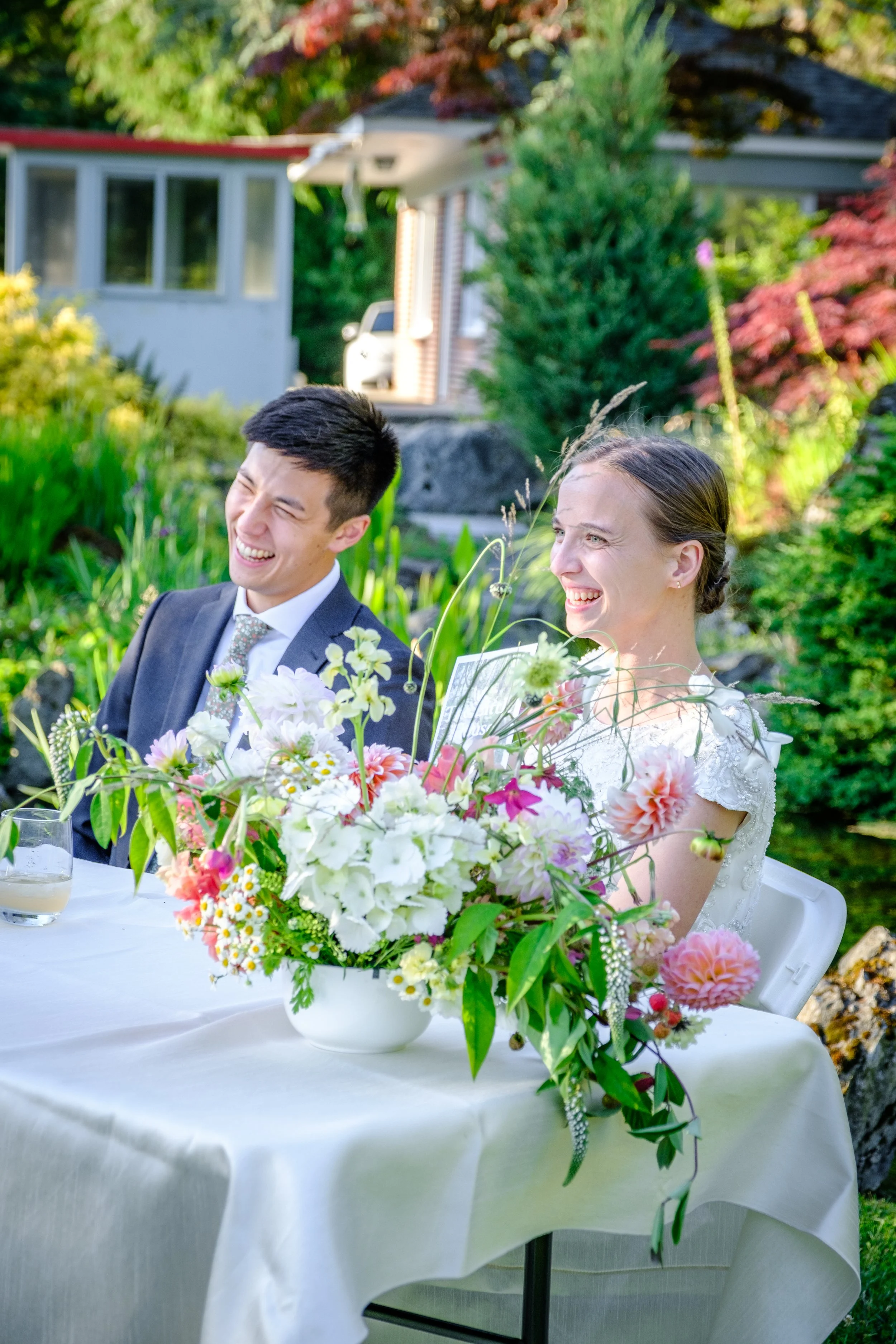 A bride and groom sitting at a table outdoors, smiling and laughing, with a large bouquet of colorful flowers on the table.
