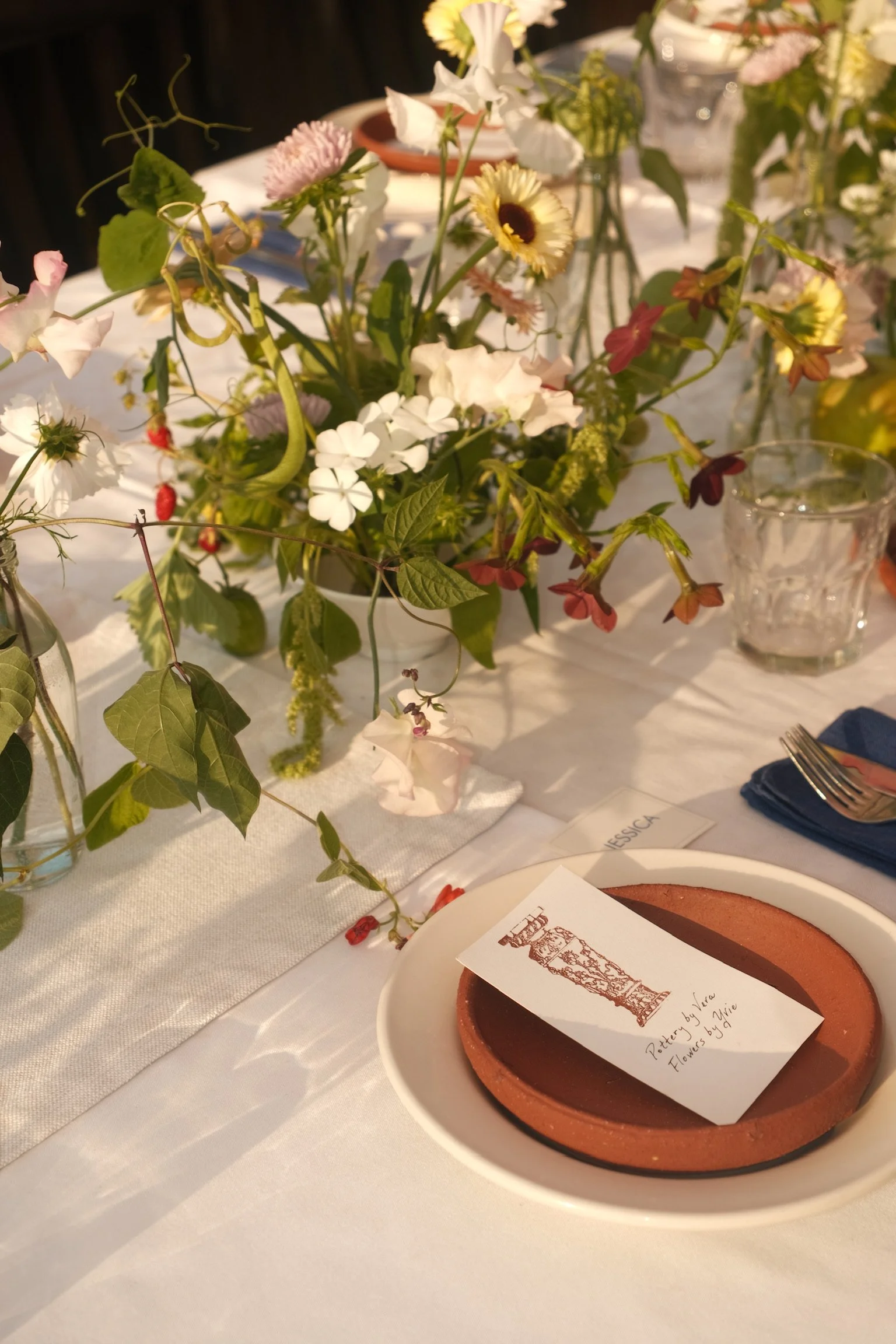 Table setting featuring a floral centerpiece, a white plate with a brown terracotta pot, and a hand-written note indicating pottery by Vera and flowers by Nikkie.