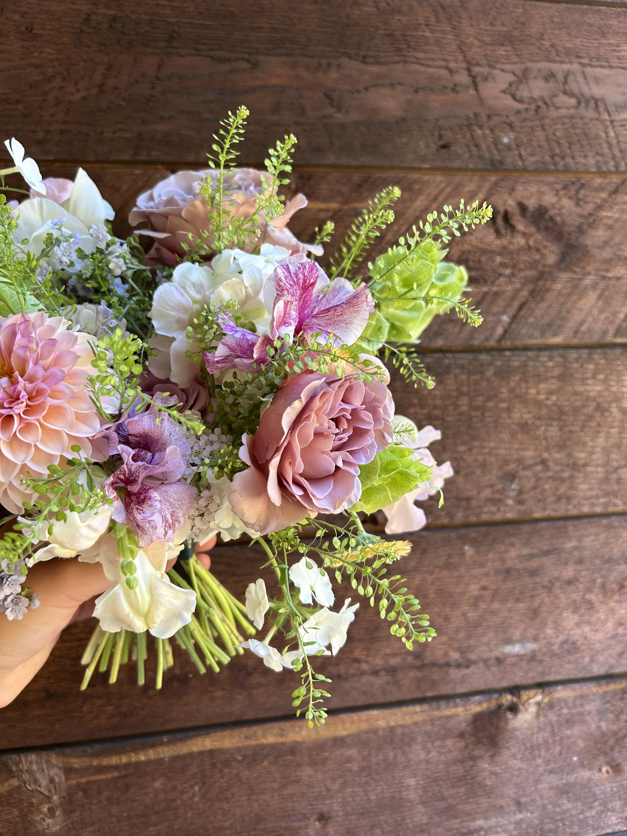 Hand holding a bouquet of mixed pink, white, purple, and green flowers against a wooden background.