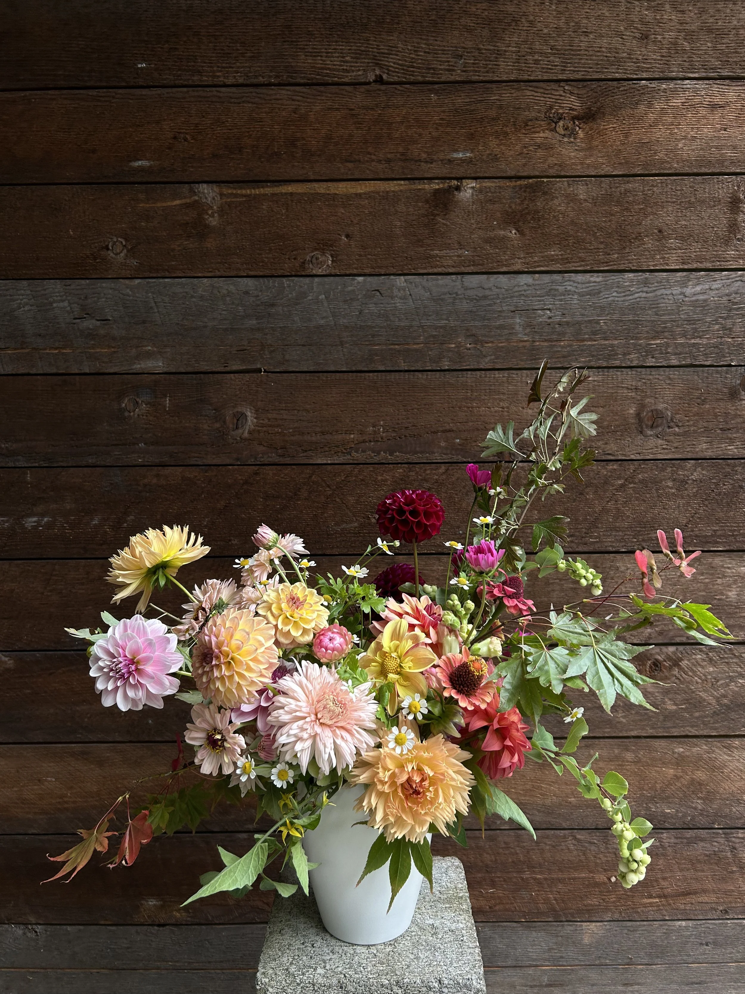 A bouquet of various colorful flowers in a white vase on a stone surface against a wooden wall background.