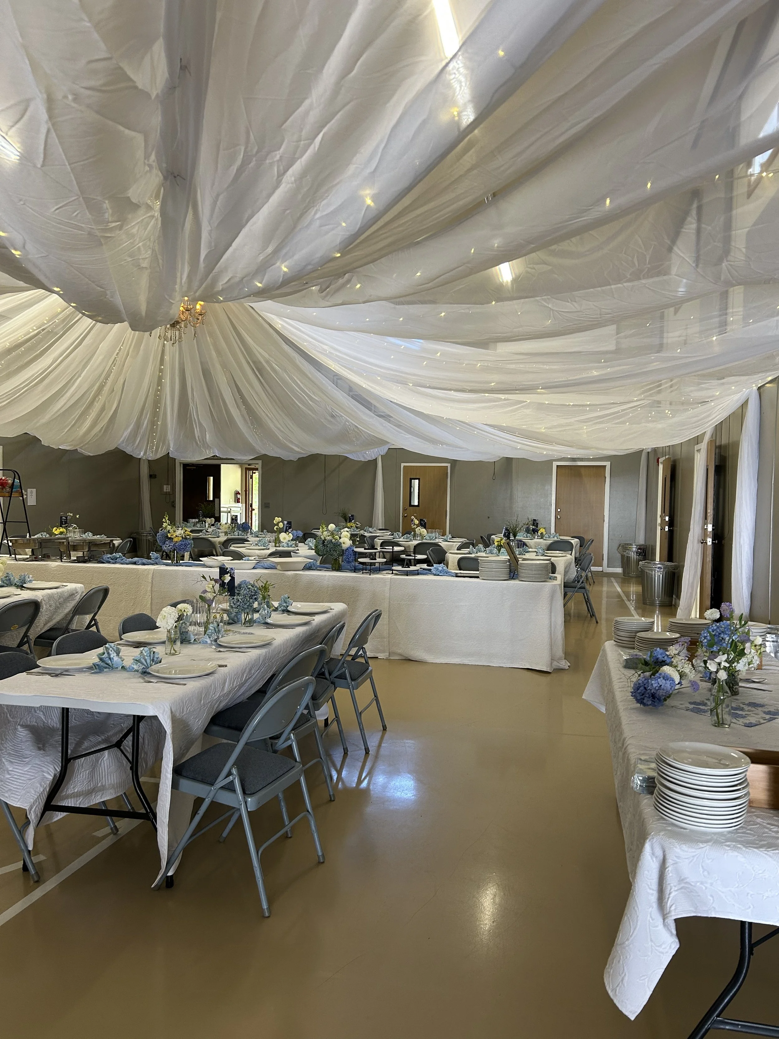 A banquet hall decorated with white drapes on the ceiling, long tables with white tablecloths, and floral centerpieces in shades of white, blue, and purple. Plates and stacks of dishes are set up on side tables.