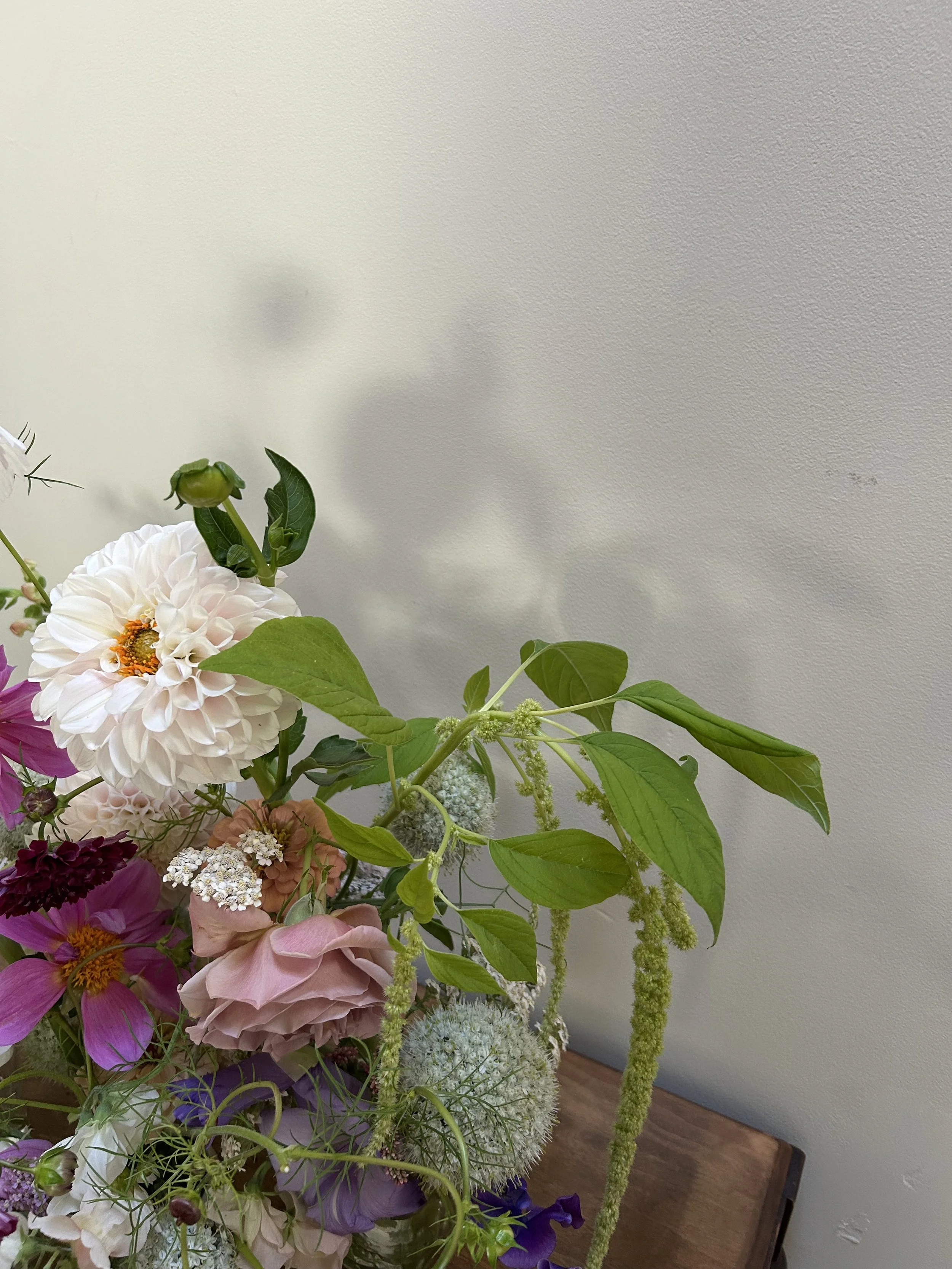 A floral arrangement with white, pink, purple, and green flowers and foliage on a wooden surface against a plain light-colored wall.