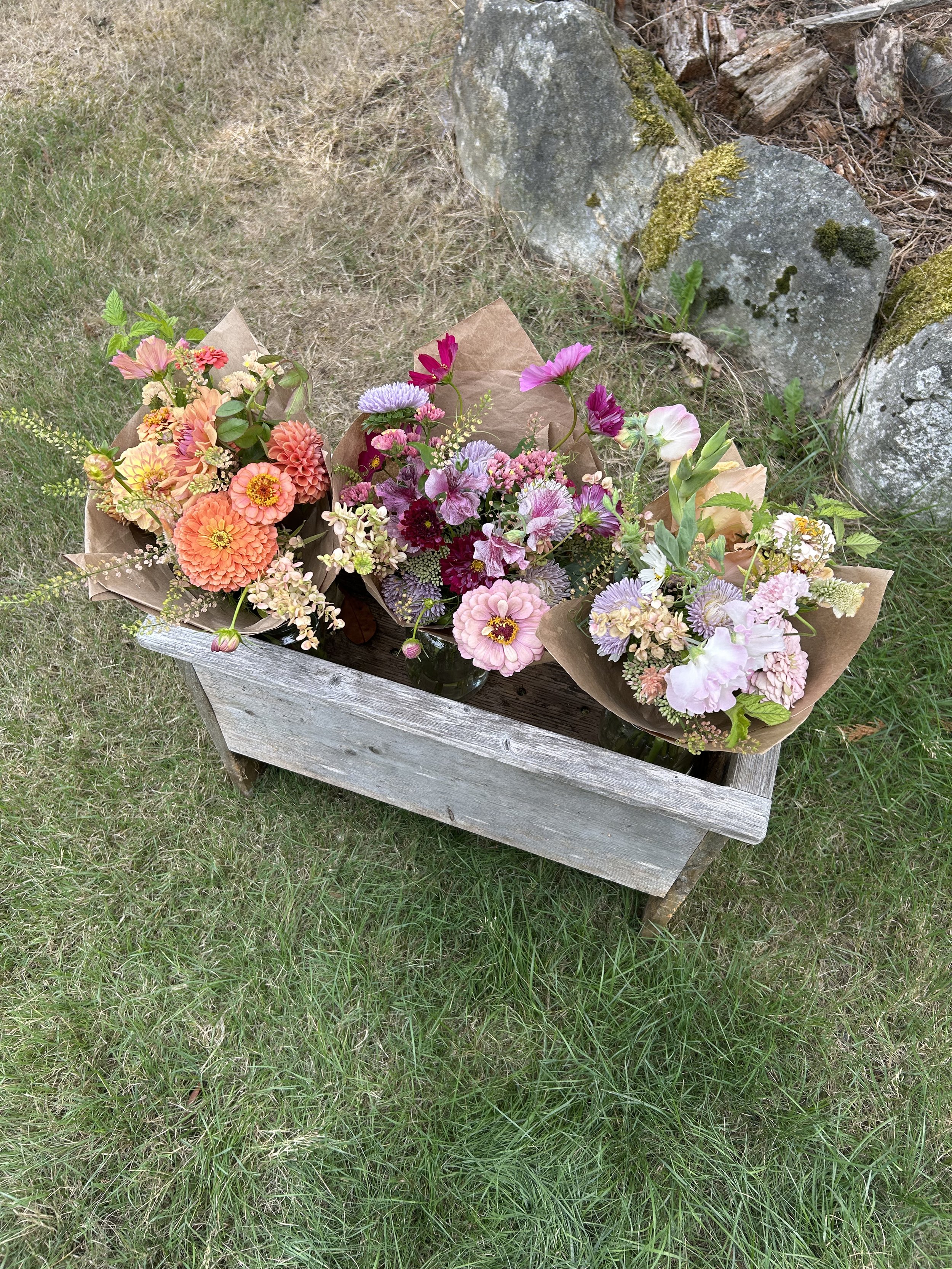 Three colorful bouquets of flowers wrapped in brown paper, arranged in glass jars, on a weathered wooden table outdoors with grass and rocks in the background.