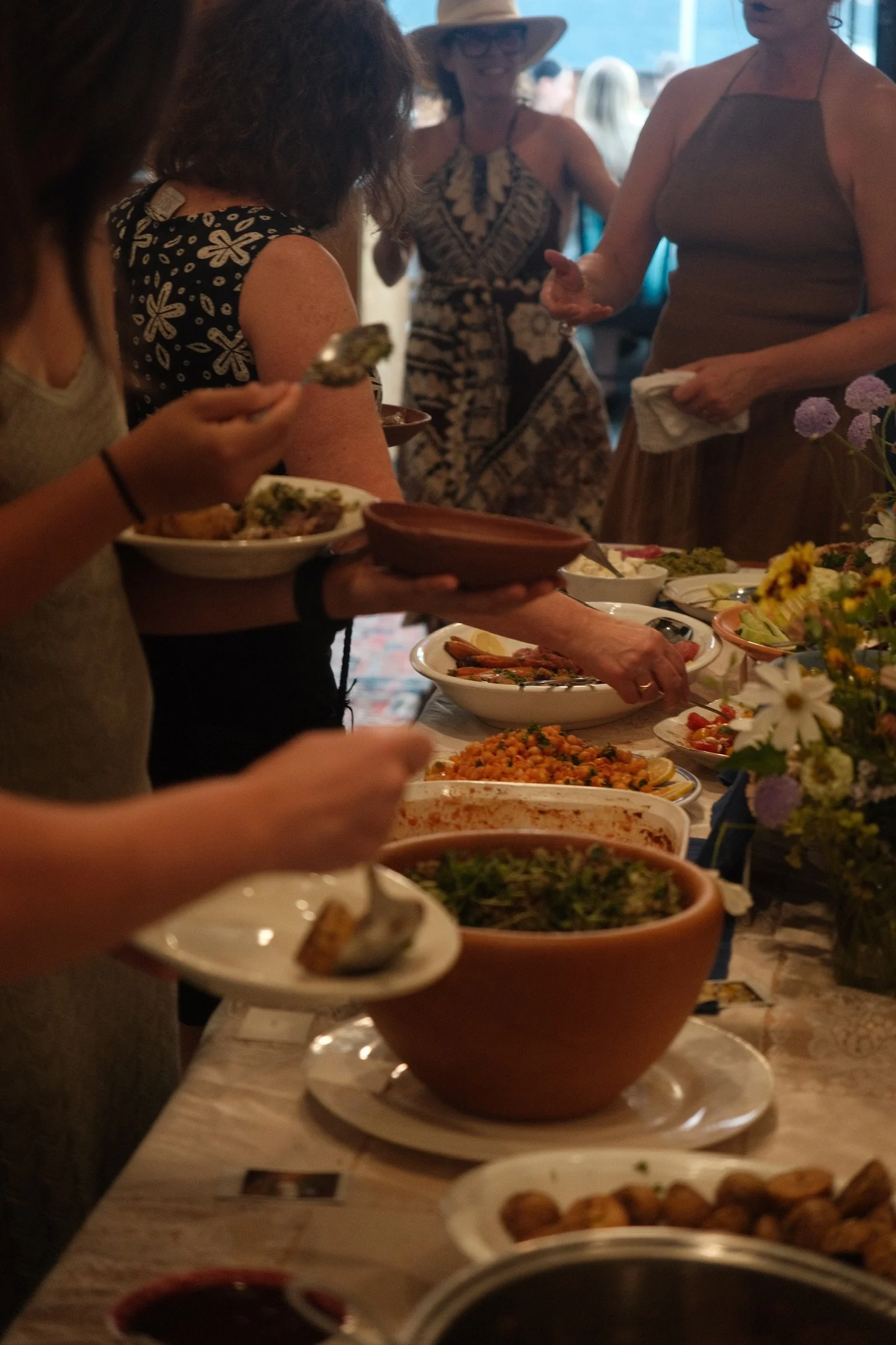 People serving and eating food at a buffet-style gathering, with dishes, bowls, and flowers on the table.