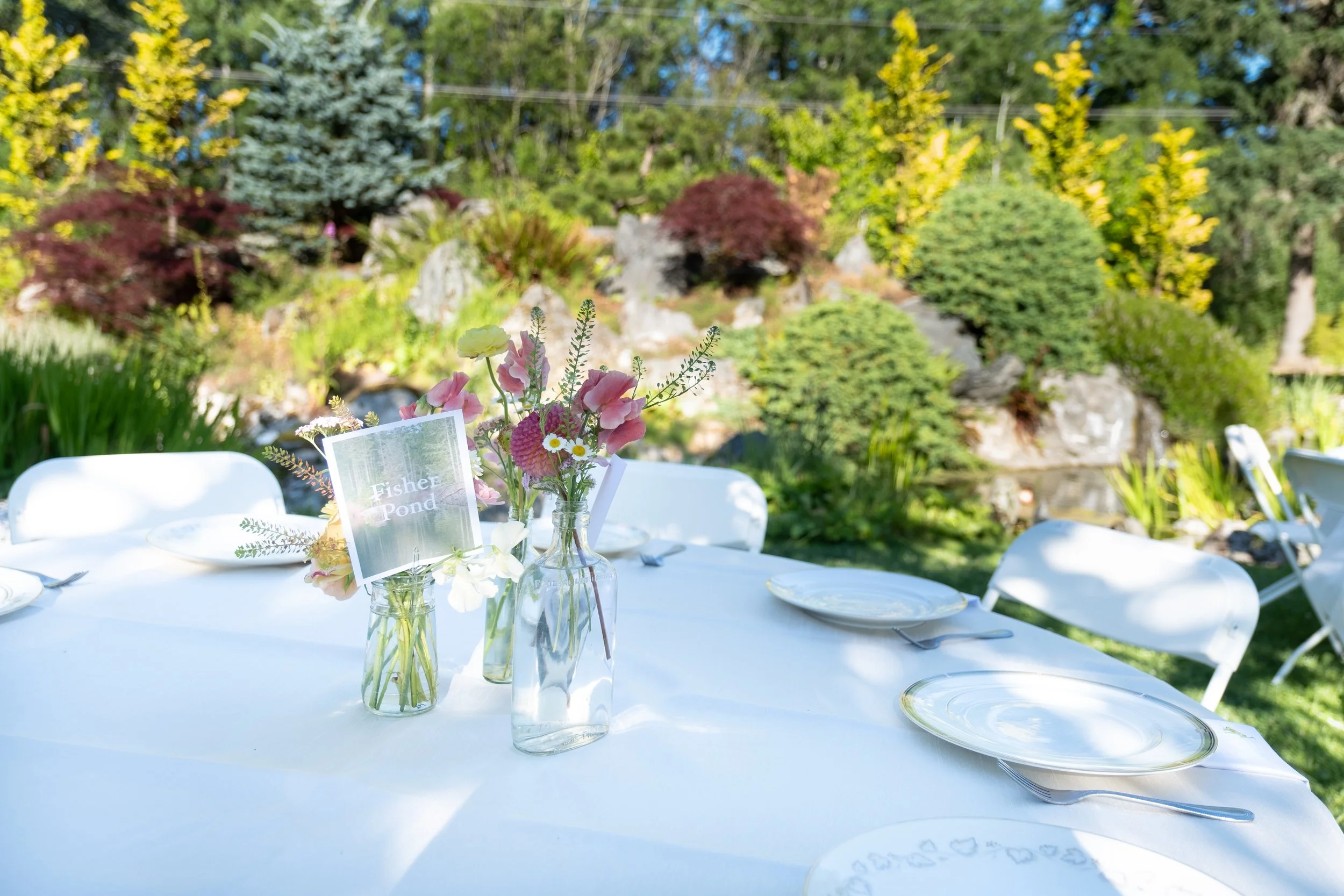 Outdoor table set for a gathering with white tablecloth, white plates, silverware, and floral centerpieces in glass bottles, in a garden with a pond and lush greenery.