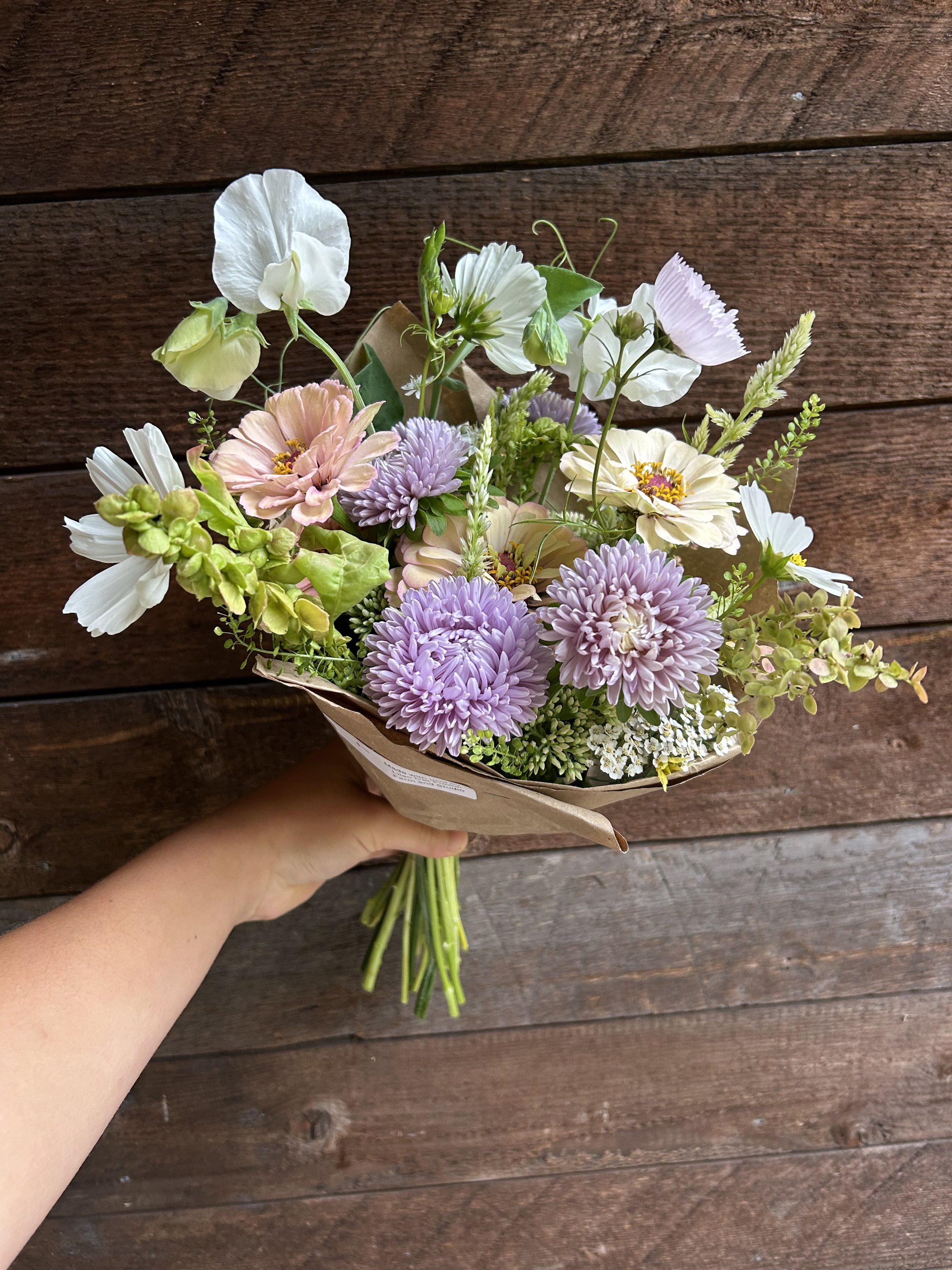 A person holding a bouquet of pastel-colored flowers against a wooden background.