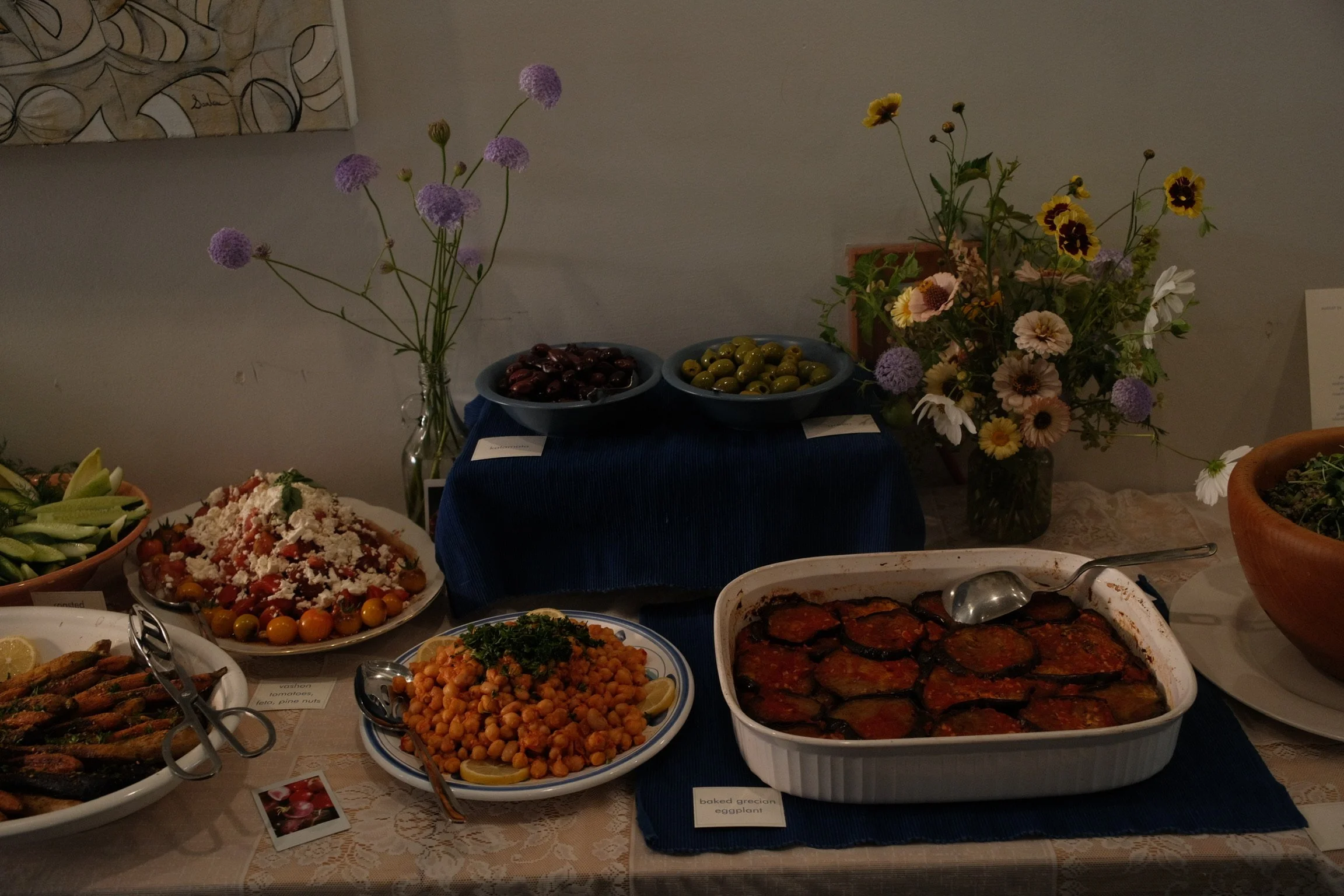 A table of Mediterranean dishes including roasted eggplant, chickpea salad, and grilled vegetables, with flower arrangements and bowls of olives.