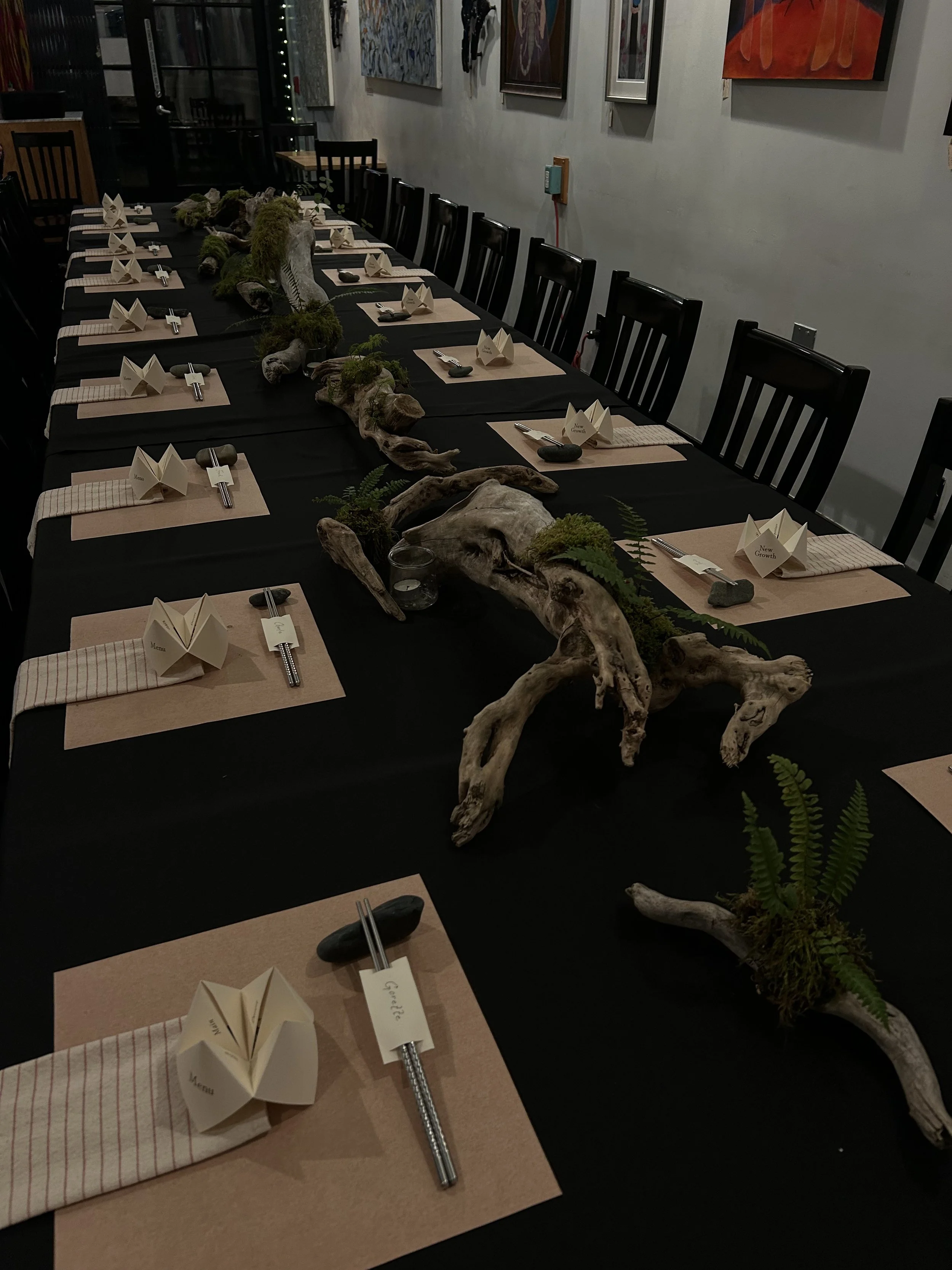 Long dining table set with placemats, folded napkins, chopsticks, and stones, decorated with driftwood, moss, and ferns for an event.