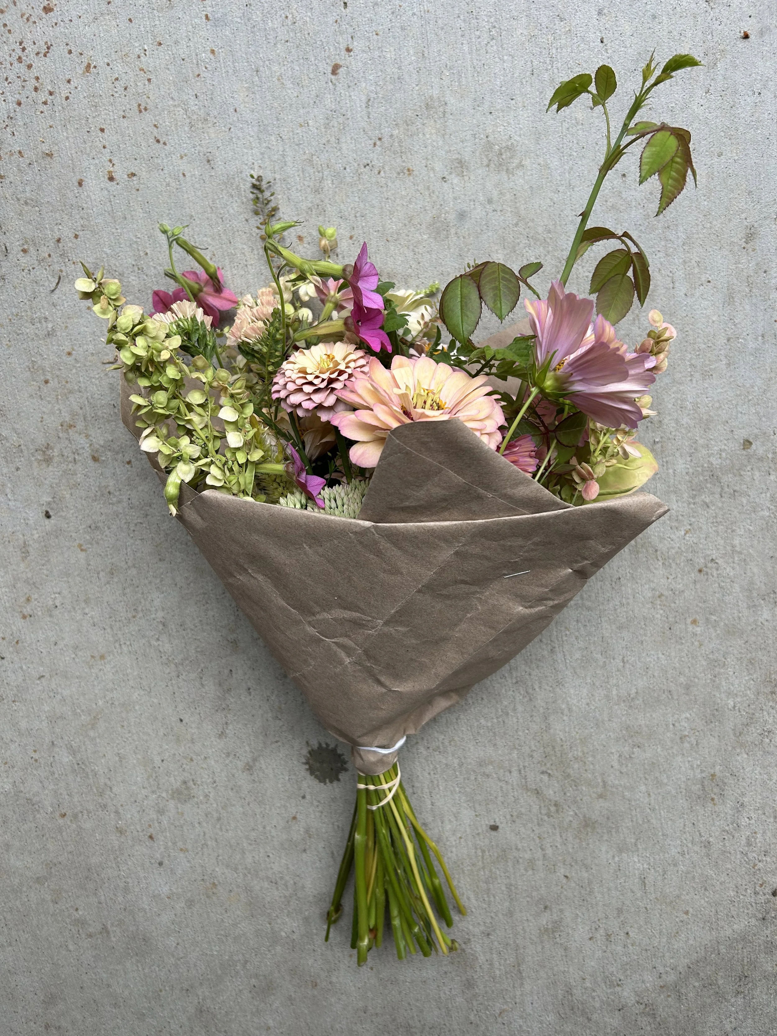 A bouquet of pink, purple, and white flowers wrapped in brown paper against a concrete background.