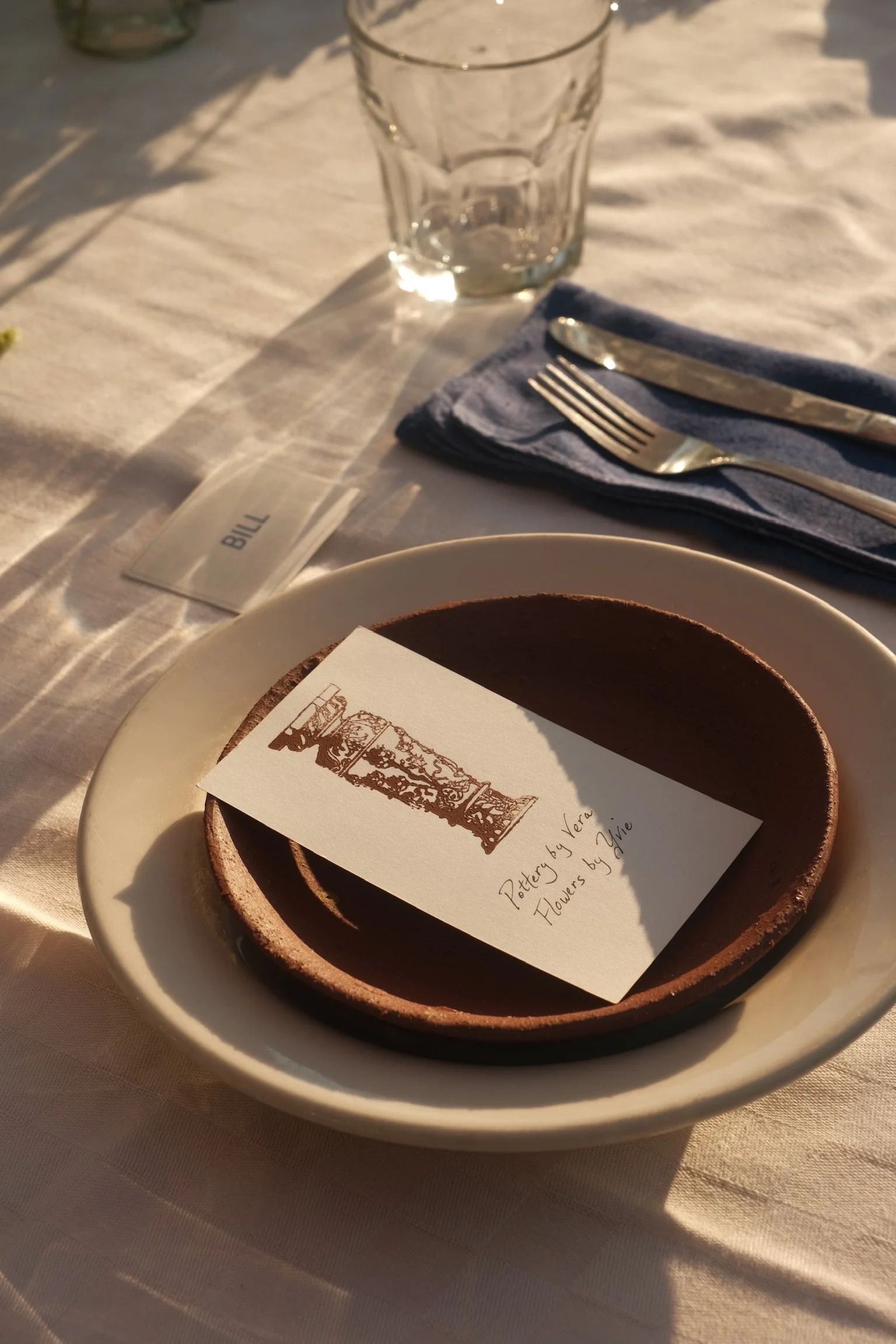 A table setting with a white tablecloth, clear glass, blue napkin, fork, and knife. On a plate, there is a small chocolate tart with a card inscribed 'Pottery by Vera. Flowers by Yvie' and a bill underneath.